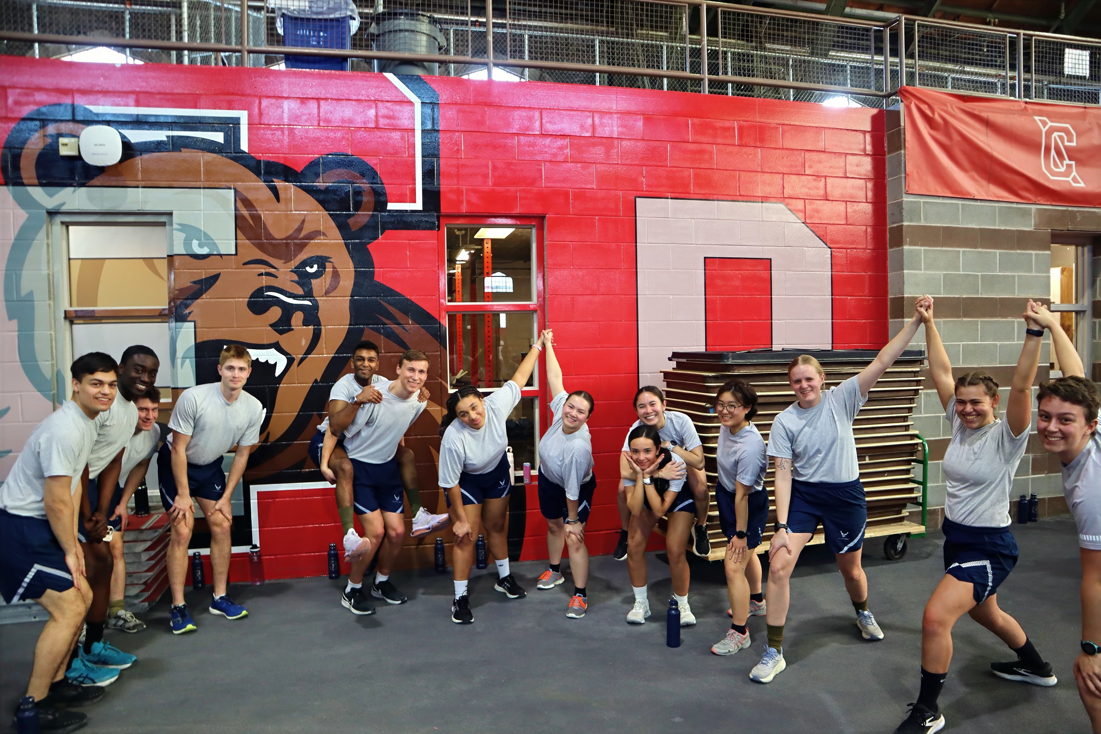 A large group of cadets in physical training gear cheering together in front of the bear mural in Barton Hall.