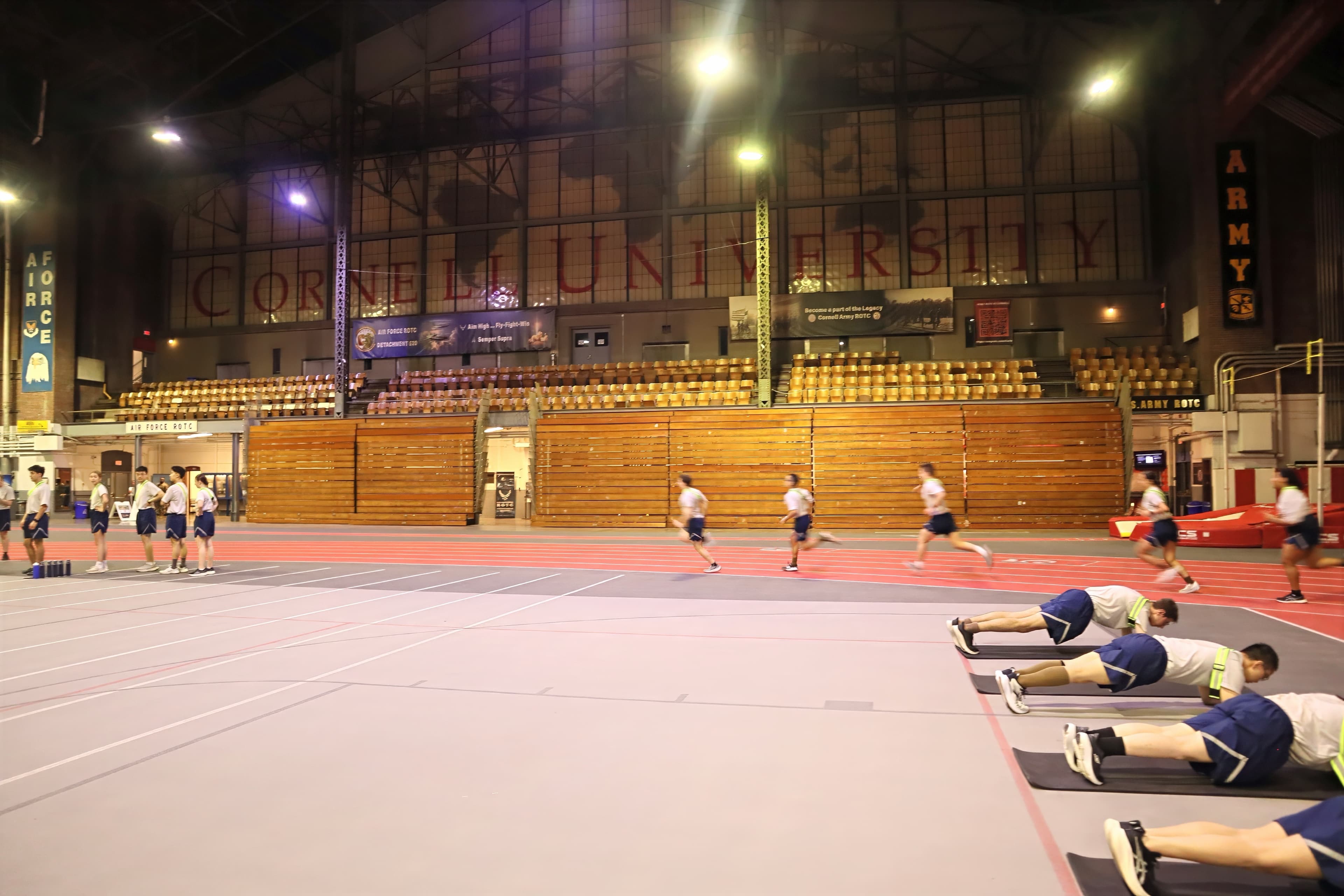 Cadets hold planks on mats while others sprint the Barton Hall track during morning physical training, beneath windows lettered Cornell University.