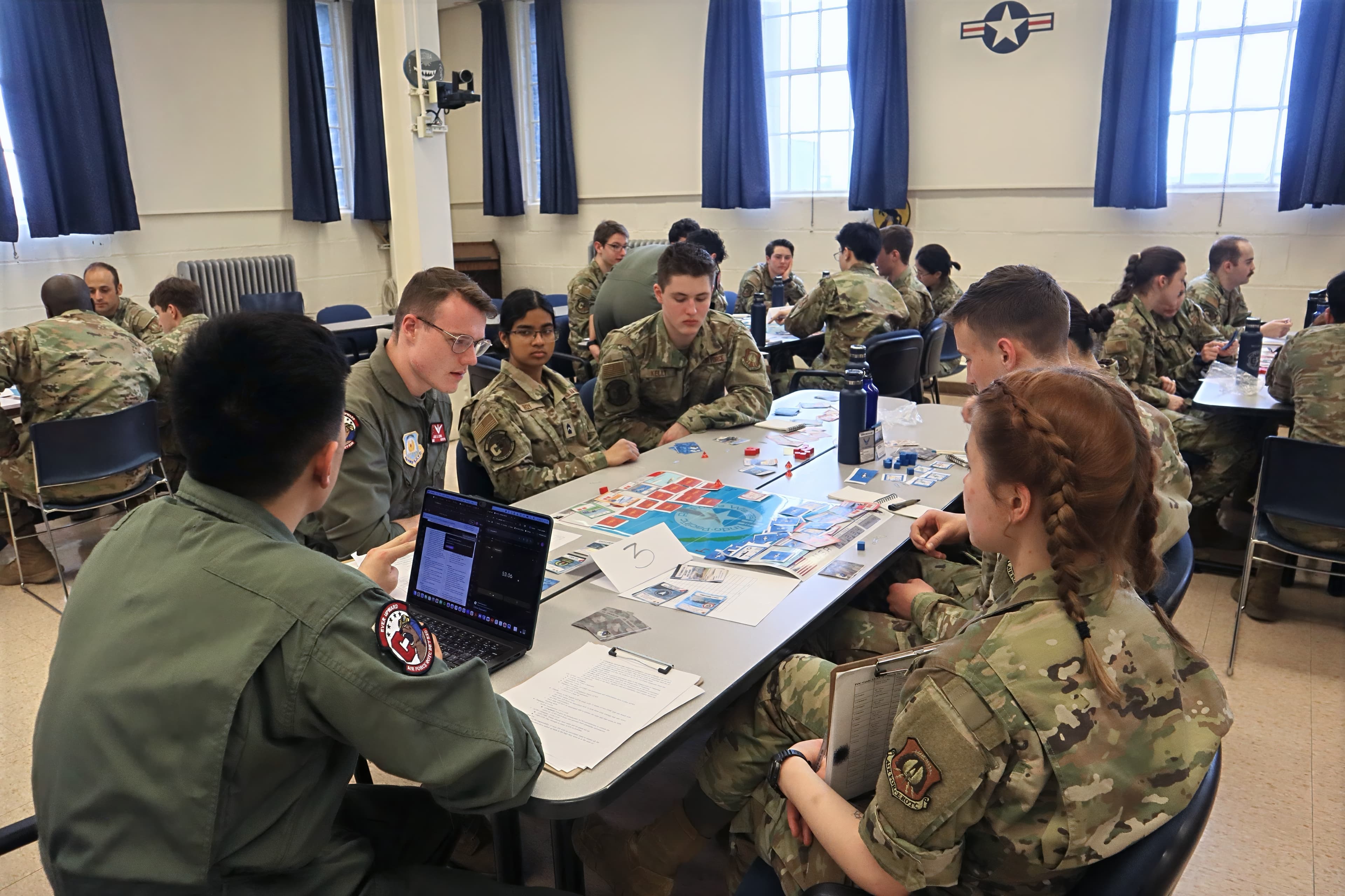 Cadre and cadets in flight suits and uniforms play a strategy wargame around a classroom table with a laptop and cards.
