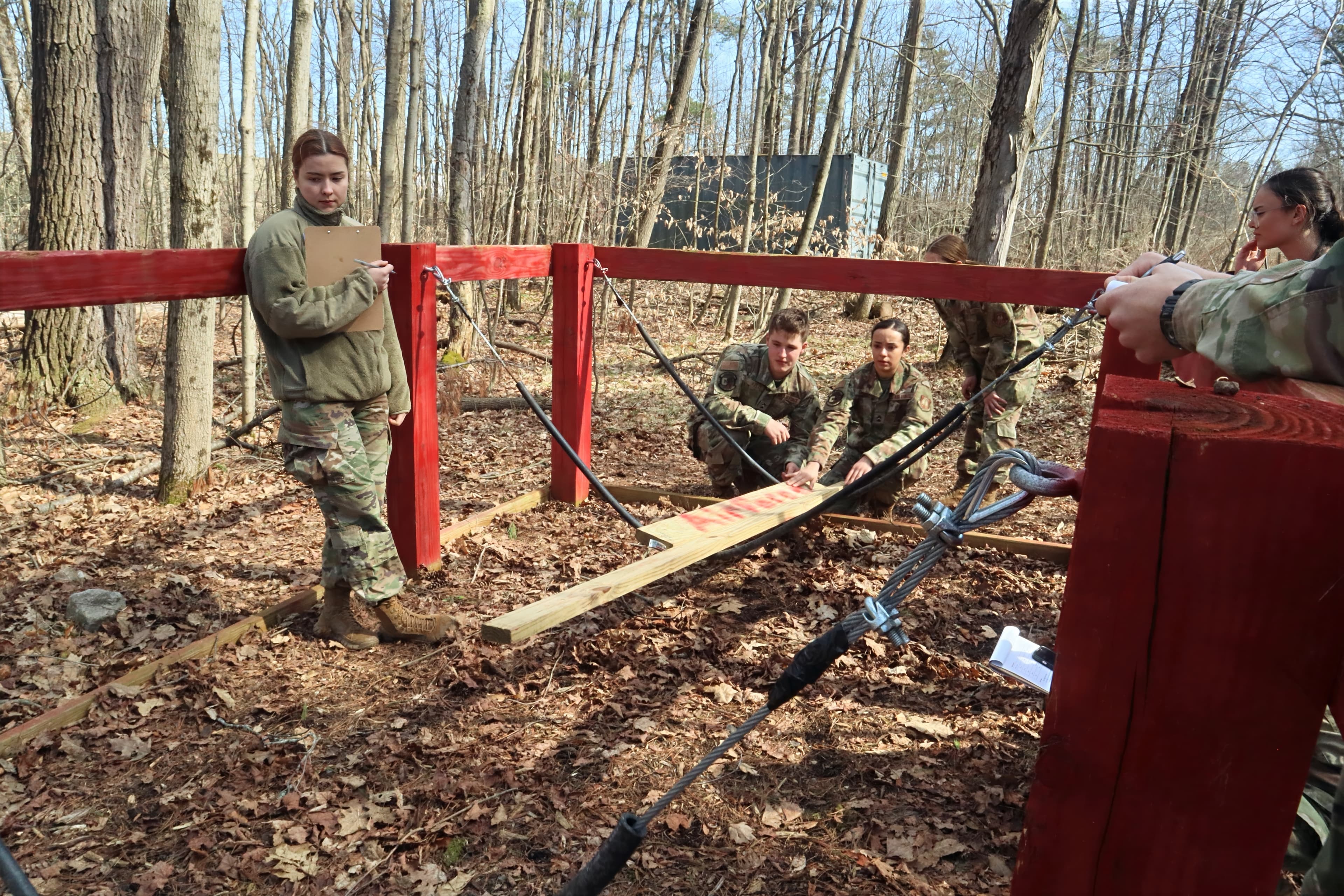 Cadets crouch around a team obstacle on the Leadership Reaction Course while an evaluator with a clipboard observes.