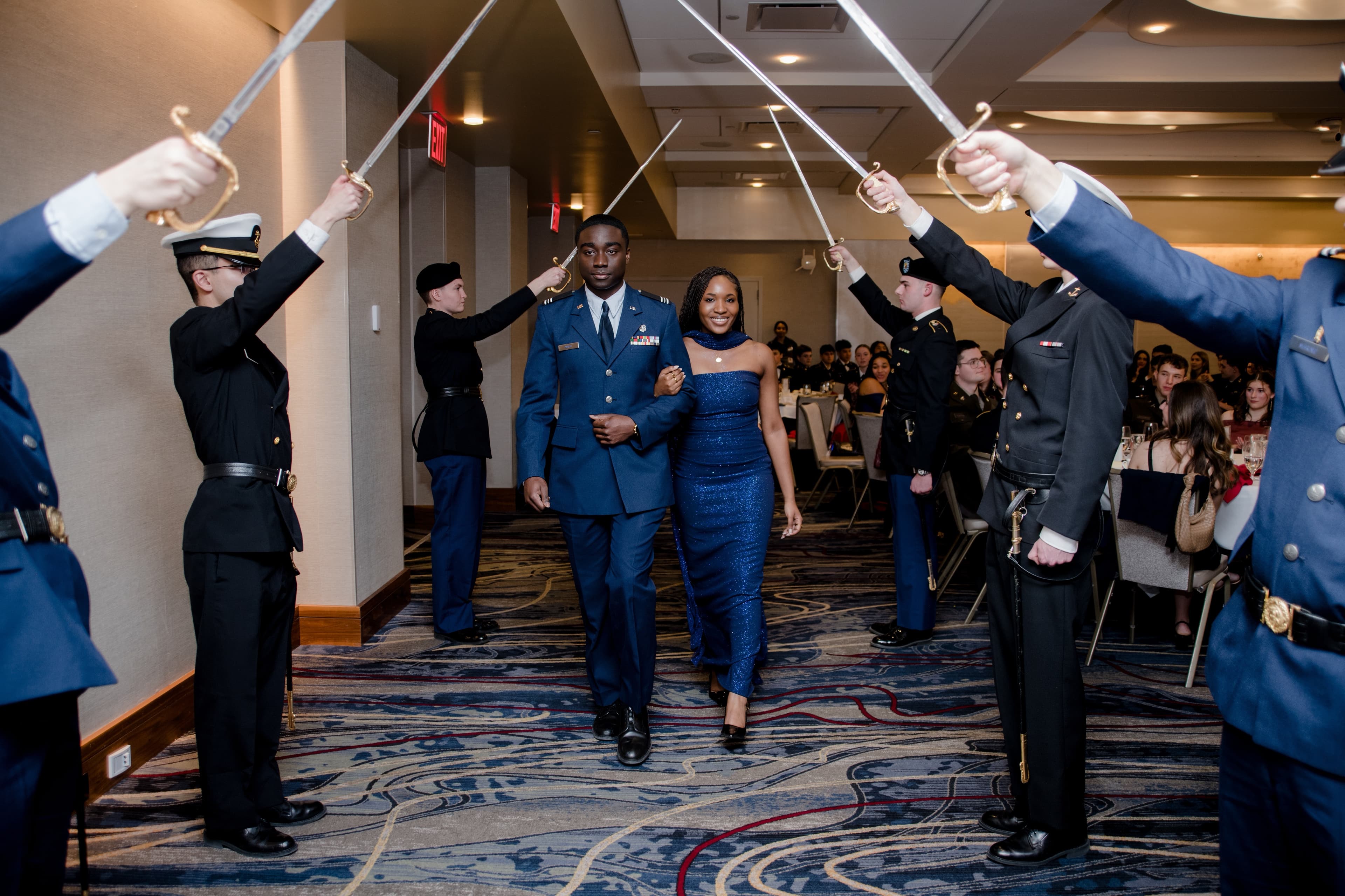 A cadet in service dress and a guest walk arm in arm through a saber arch formed by joint-service color guard members at the annual Military Ball.