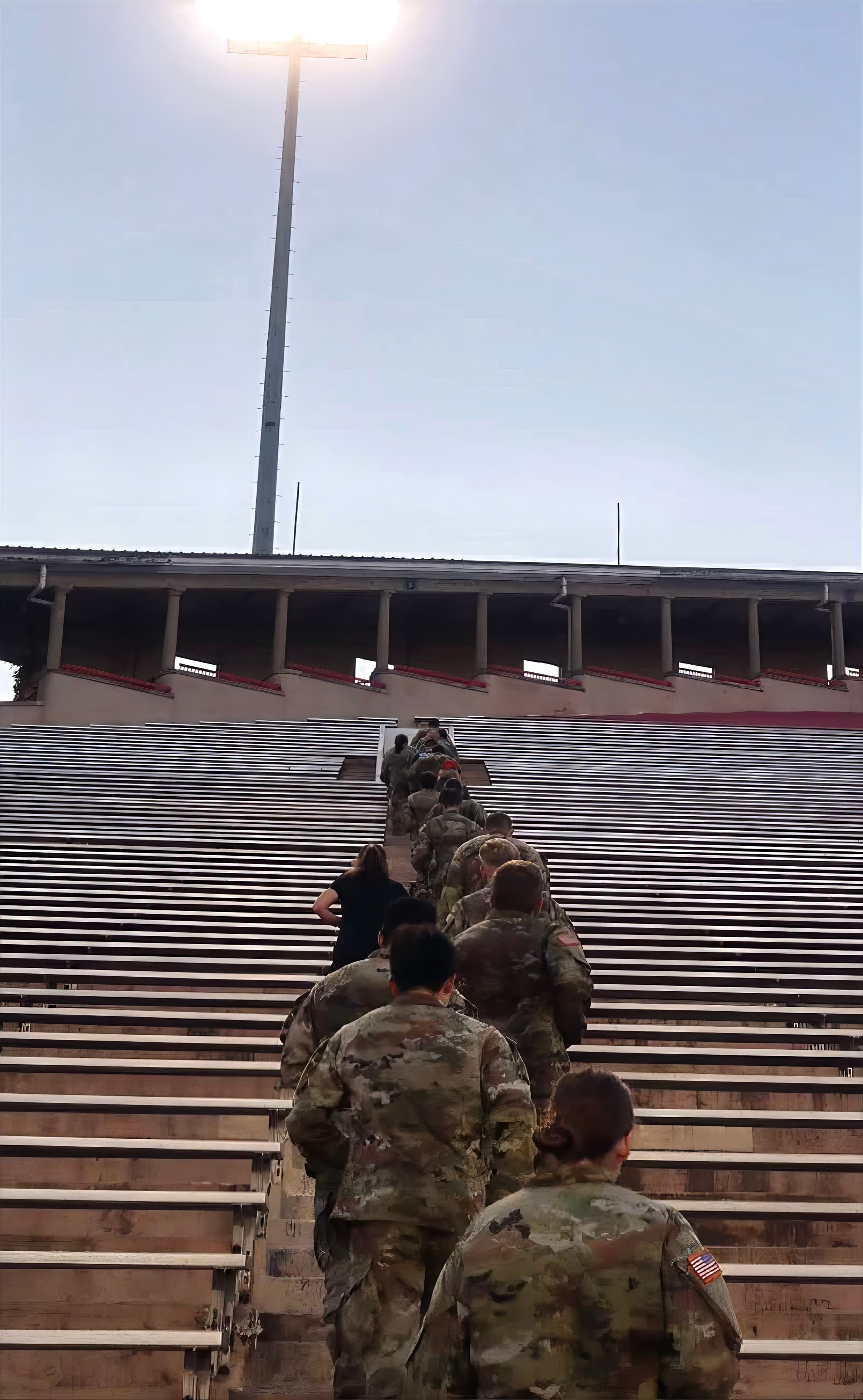 A long line of cadets runs up the stadium steps beneath a single floodlight at dawn.