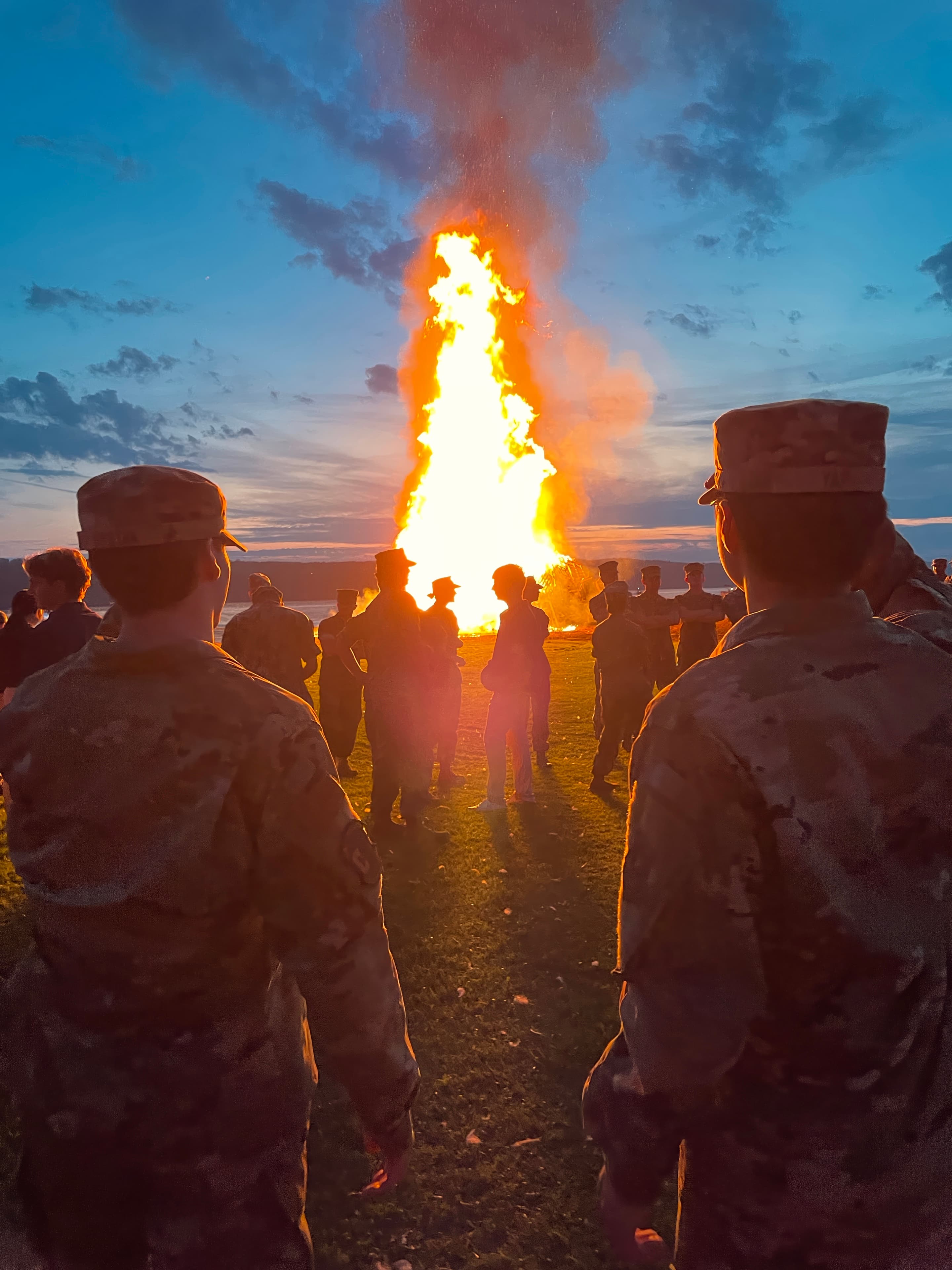 Cadets silhouetted against the burning Watchfire at dusk, with flag bearers standing by.