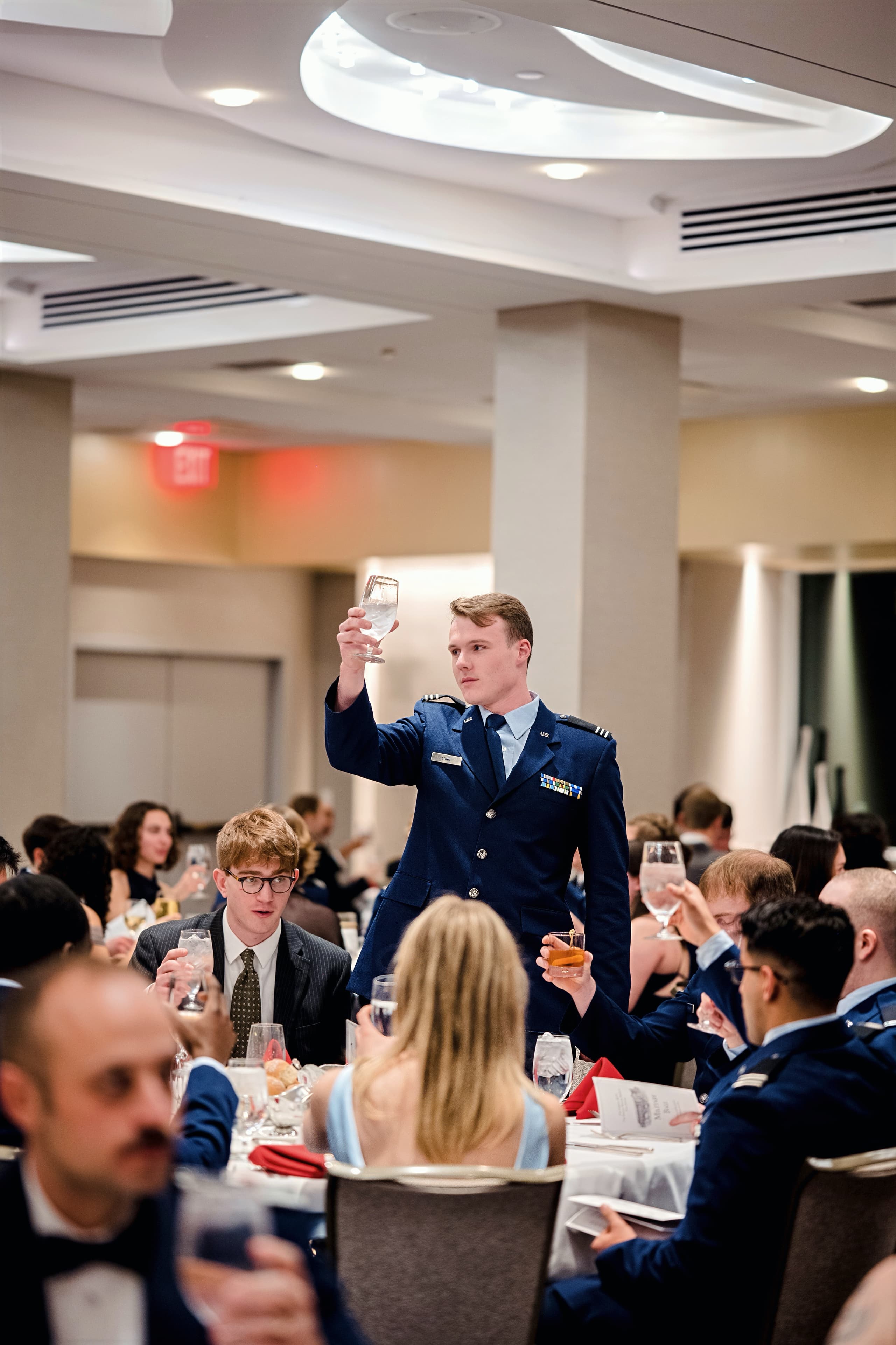 A cadet in service dress raises a glass to give a toast at a banquet table.
