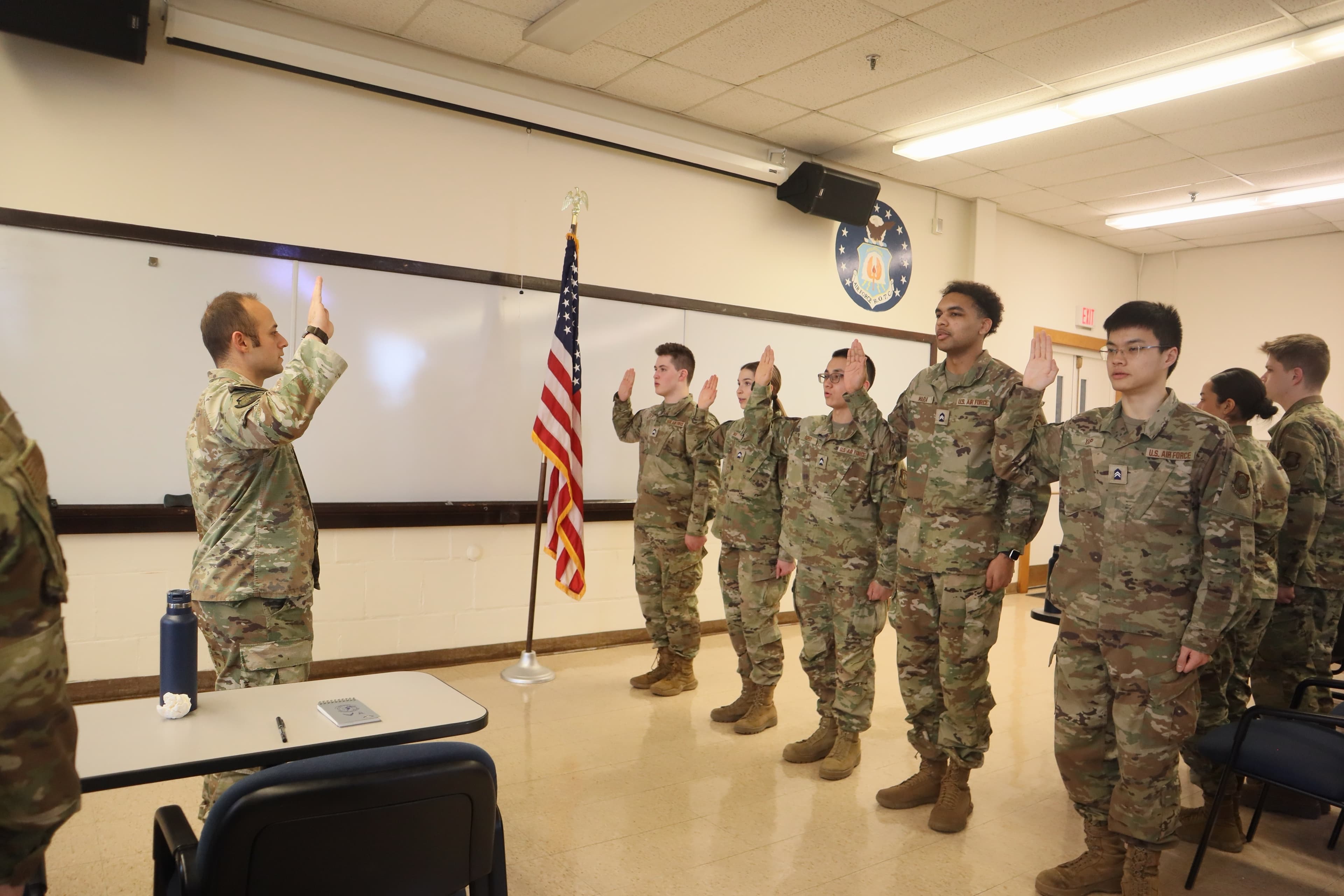 An officer administers an oath to cadets standing with their right hands raised in the detachment classroom beneath the United States flag.
