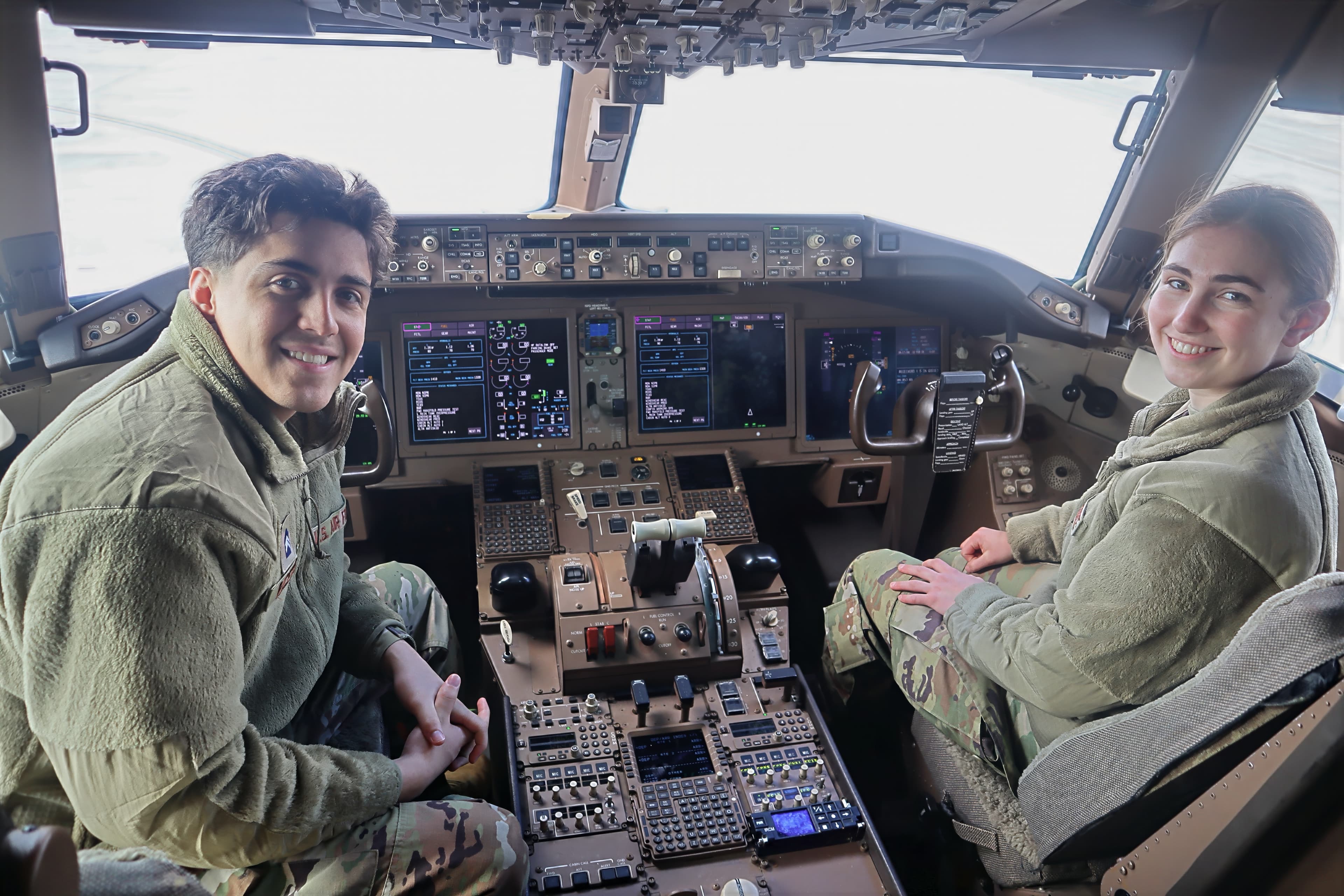 Two cadets seated at the controls in the cockpit of a large Air Force aircraft during a base visit.