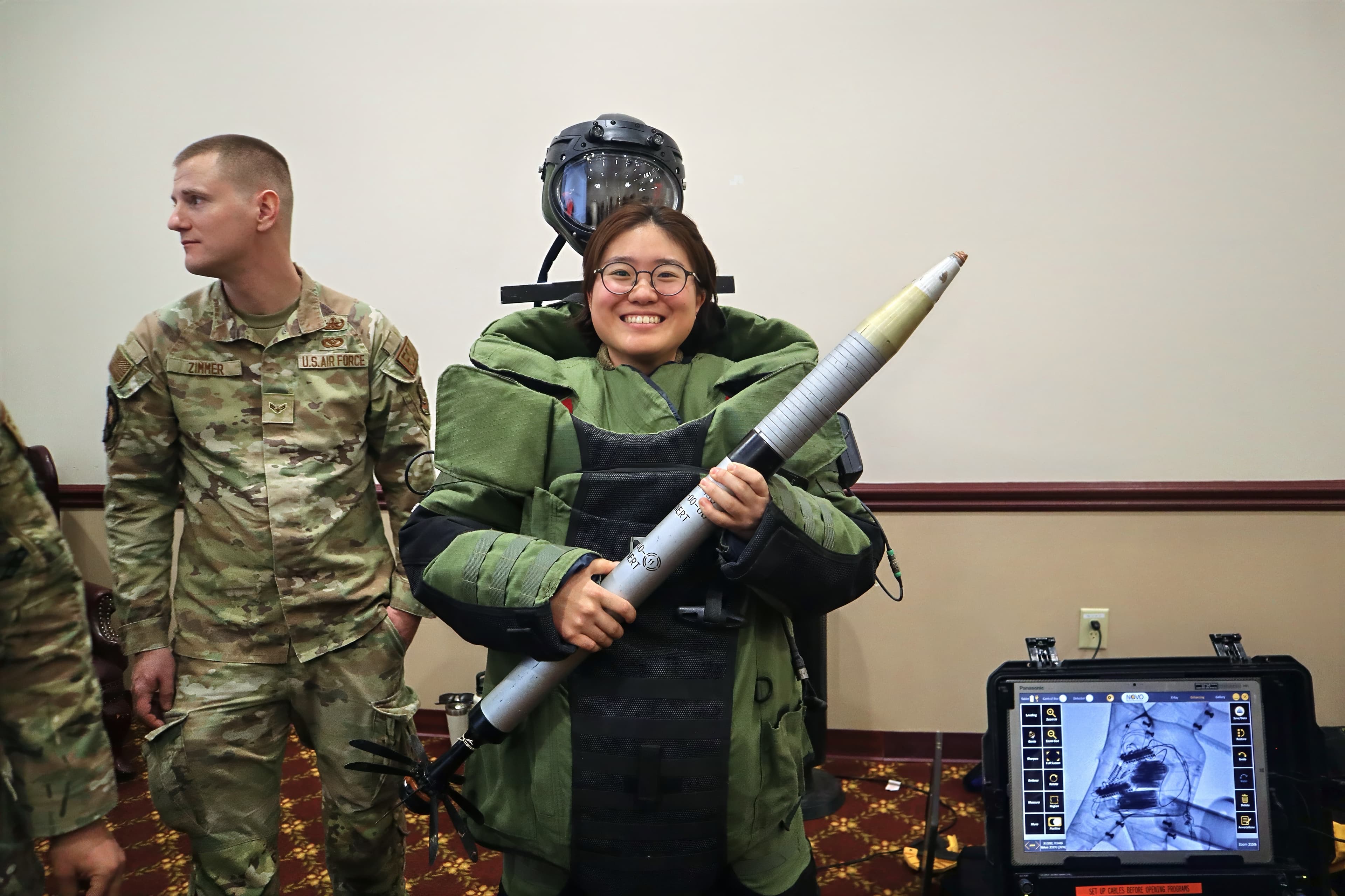 A smiling cadet wearing an explosive ordnance disposal suit holds an inert training rocket.