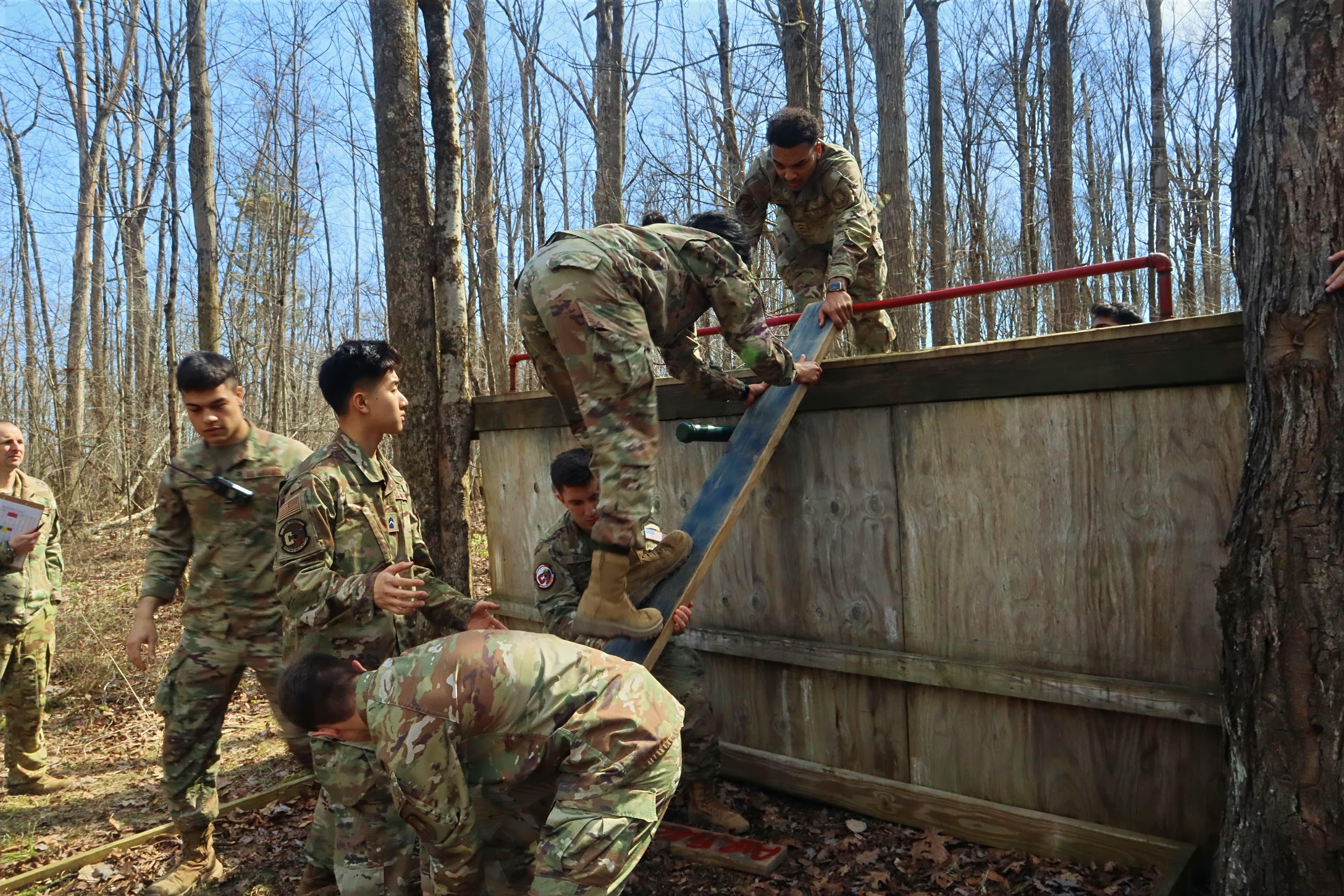 Cadets help each other over a wooden wall obstacle using a plank on the Leadership Reaction Course.