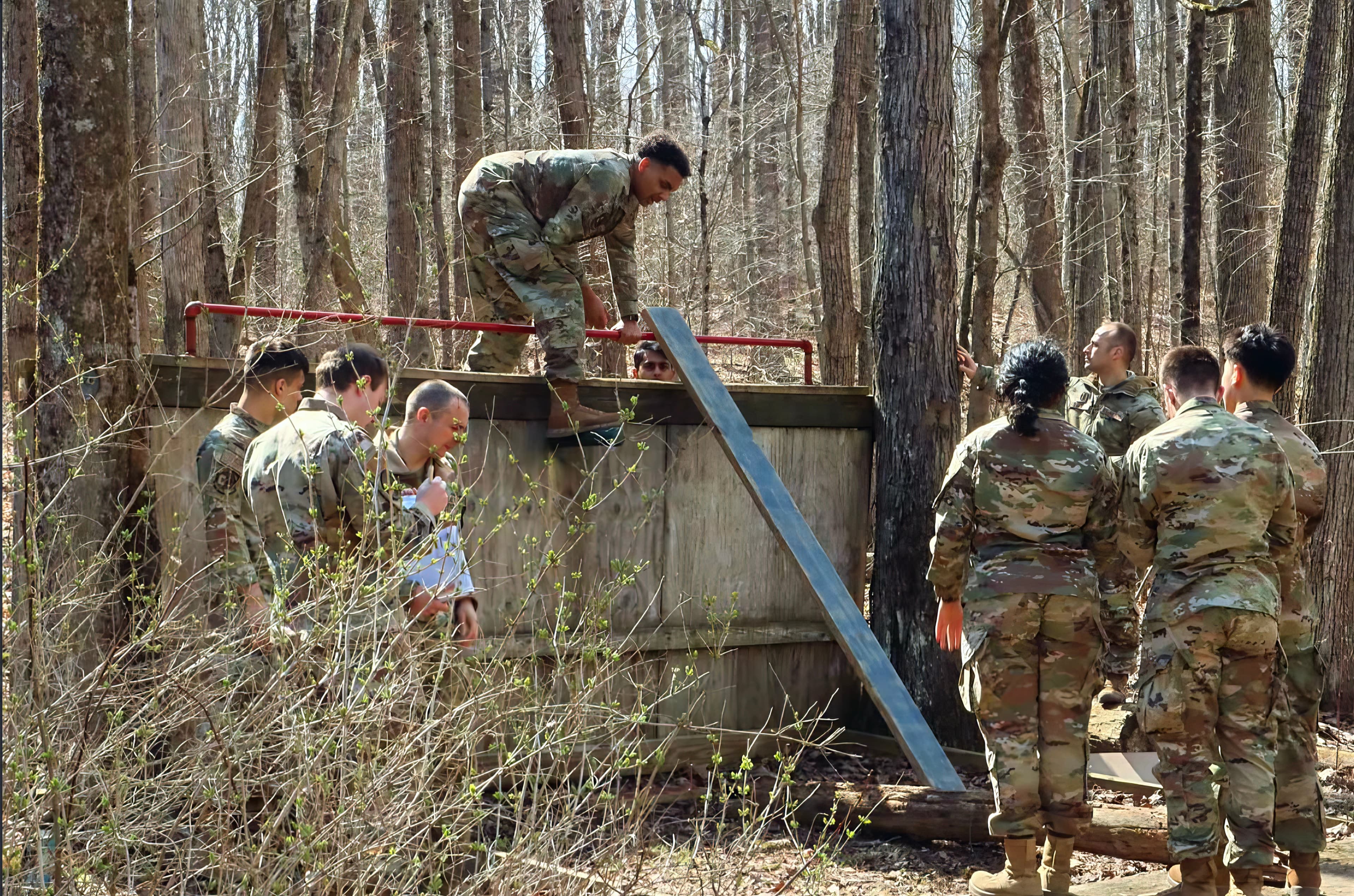 A cadet climbs a wall obstacle while teammates and an evaluator watch below.