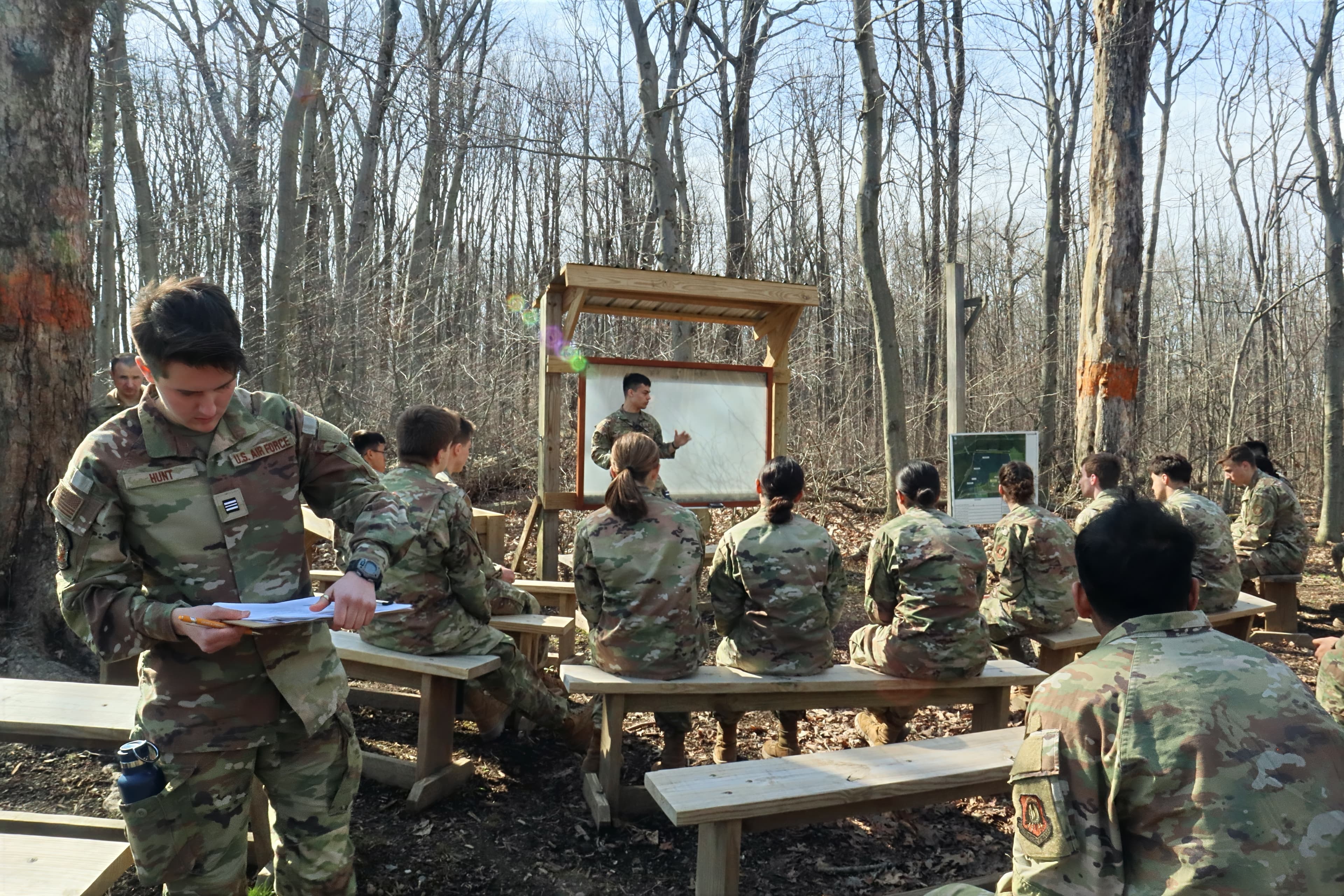 A cadet reads instructions aloud to a seated flight at an outdoor classroom among the trees.