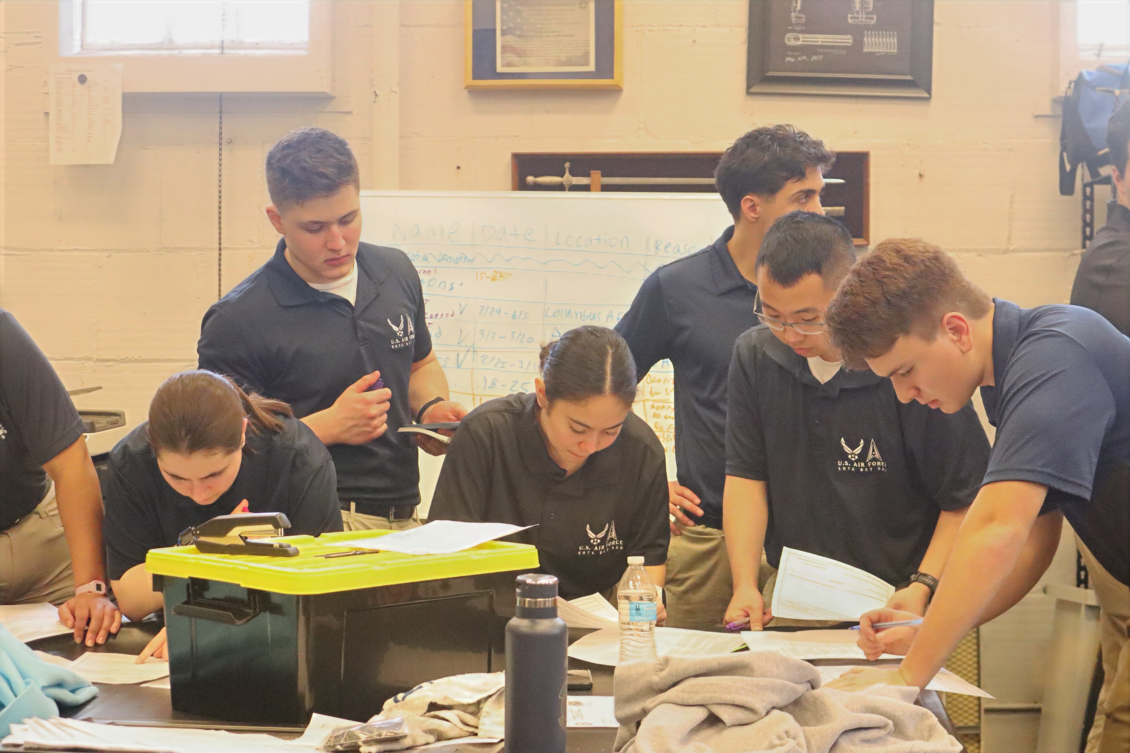 Cadets in detachment polos plan around a table stacked with papers and equipment during a group leadership problem.