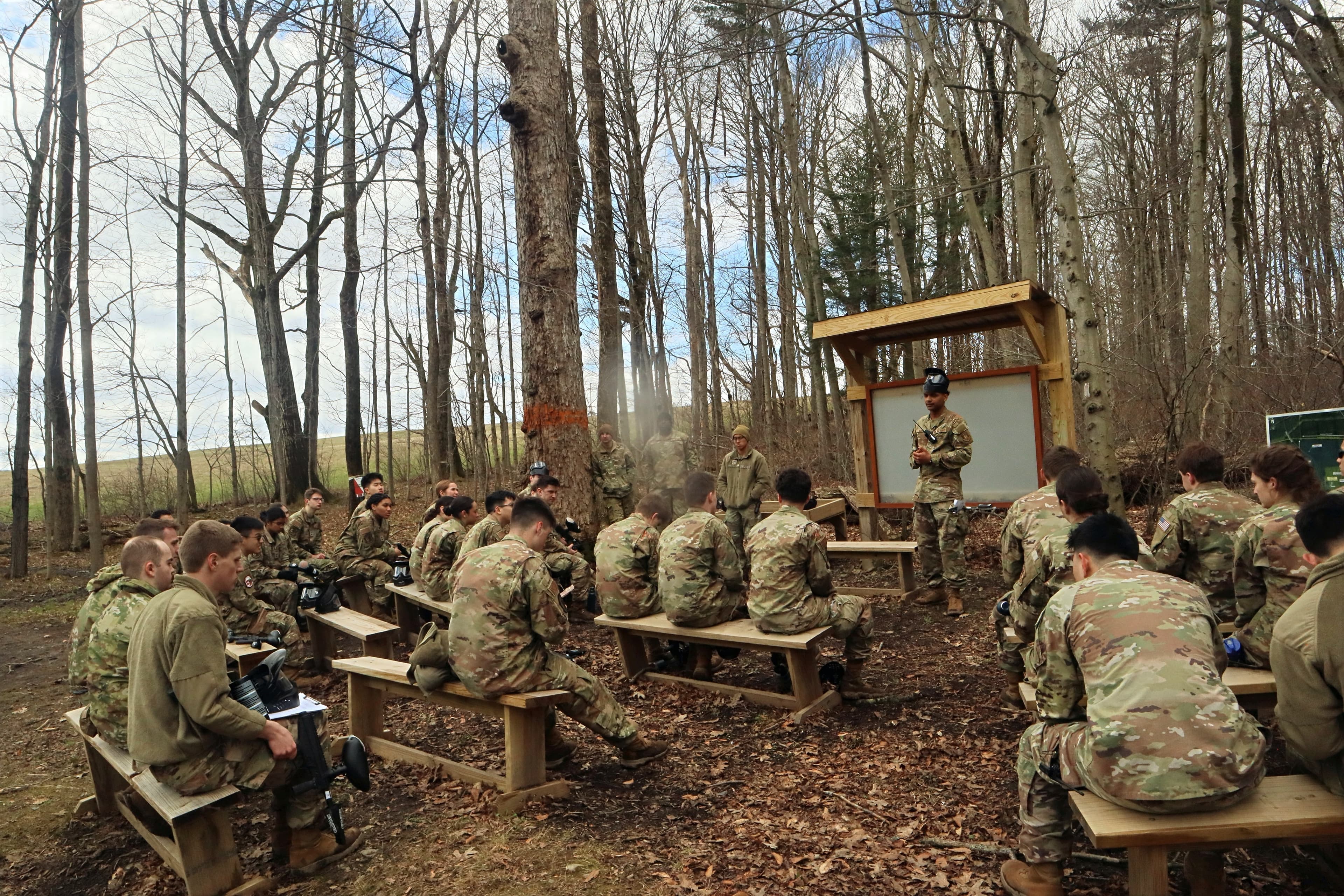 A cadet briefs a seated flight from a whiteboard at an outdoor classroom during a field training exercise.