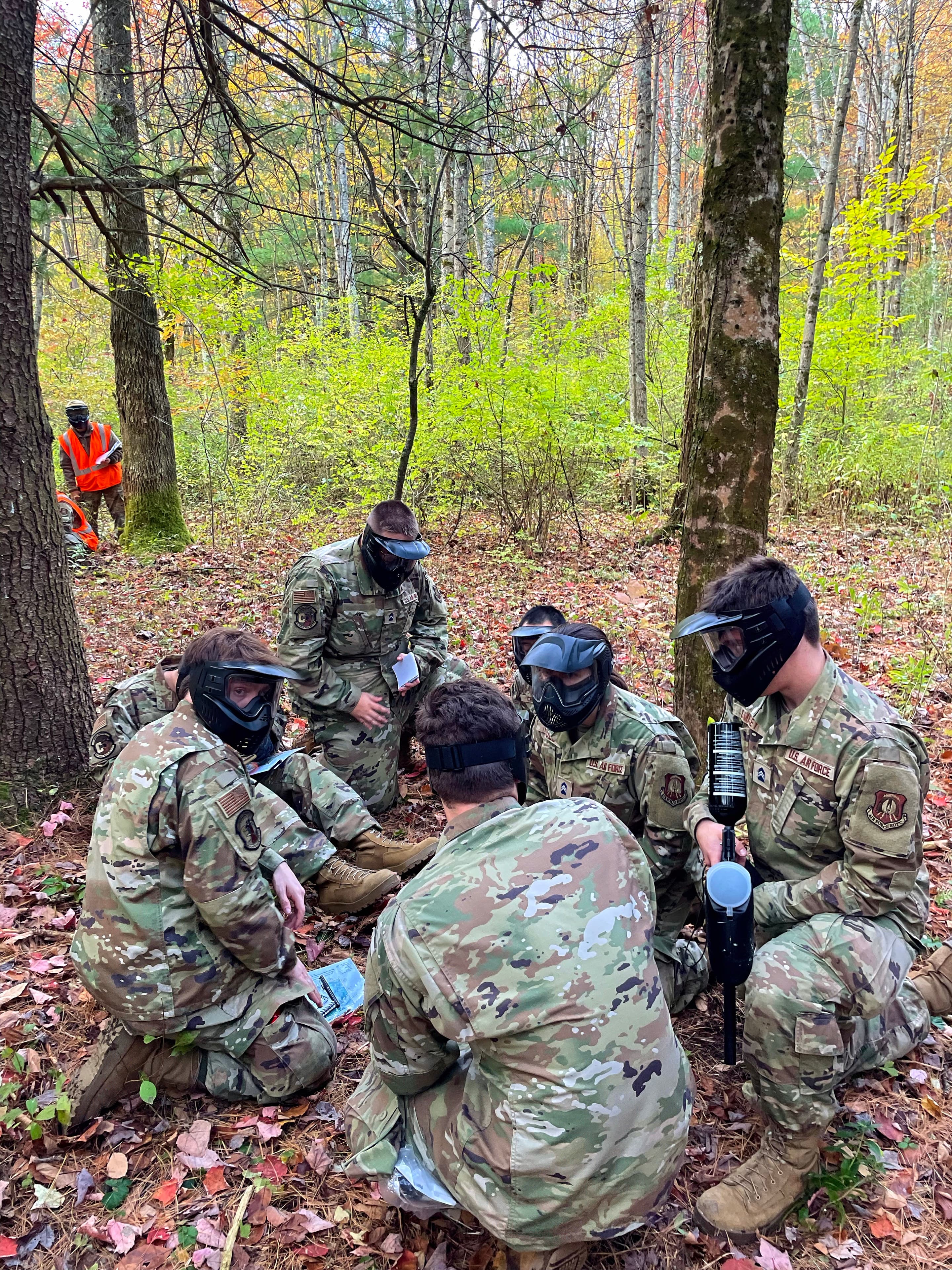 Cadets with training markers crouch in a planning circle among the trees.