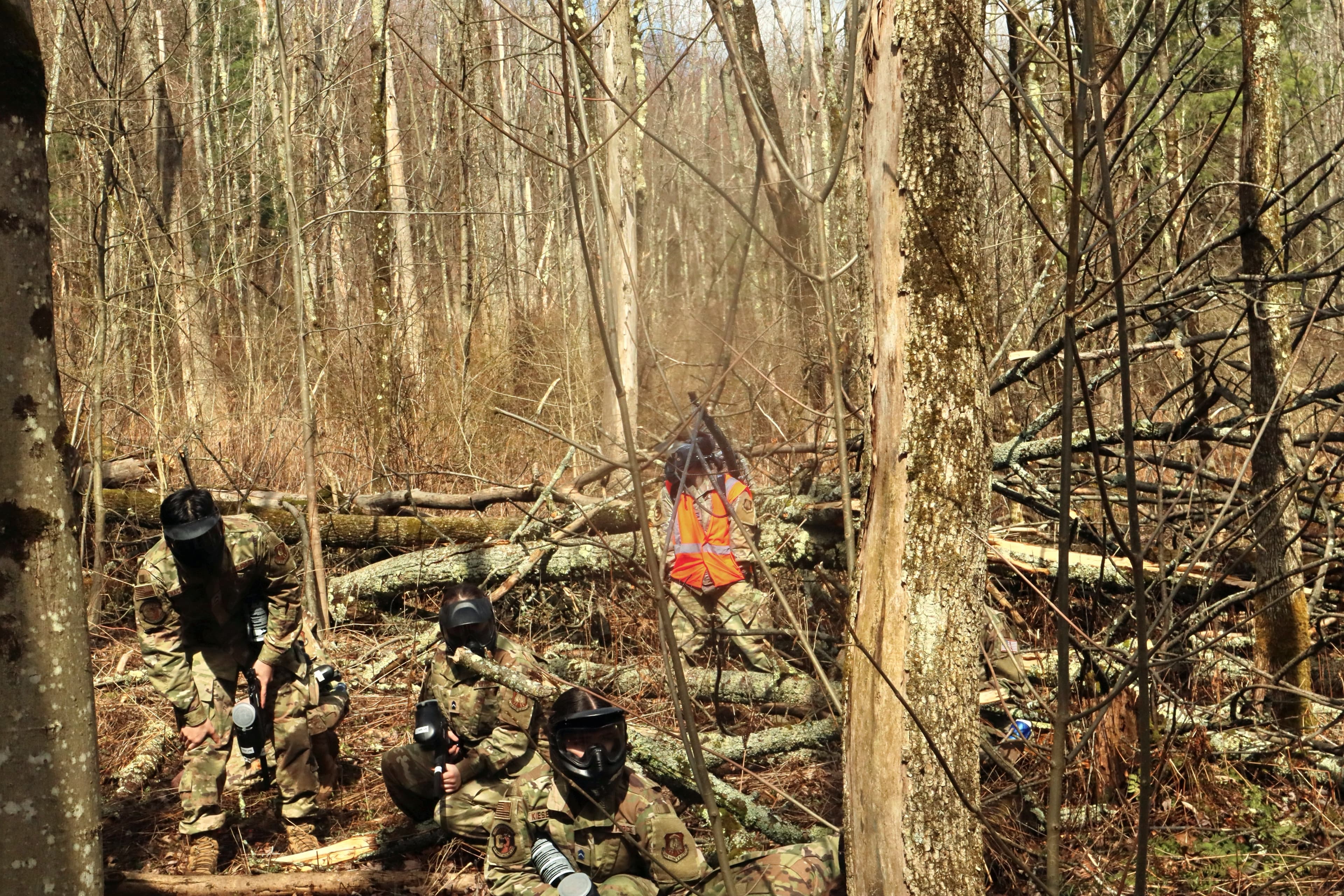 A team of cadets in masks moves low toward the camera through the woods.