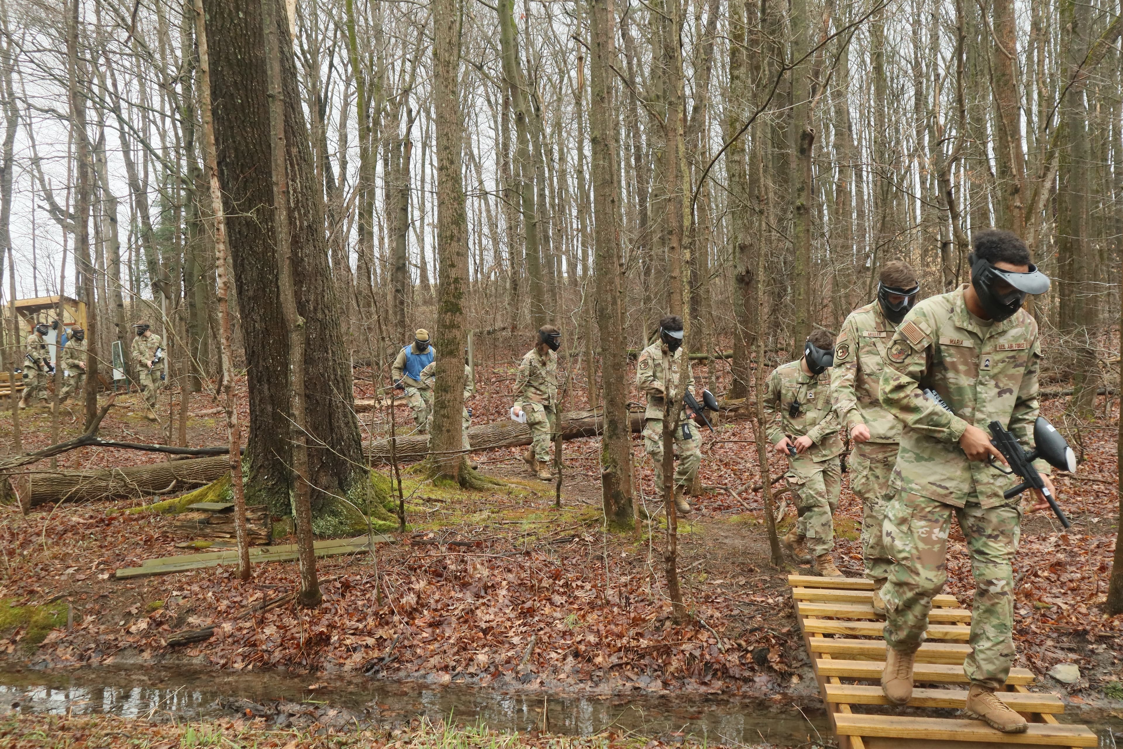 Cadets in protective masks cross a wooded path in single file.