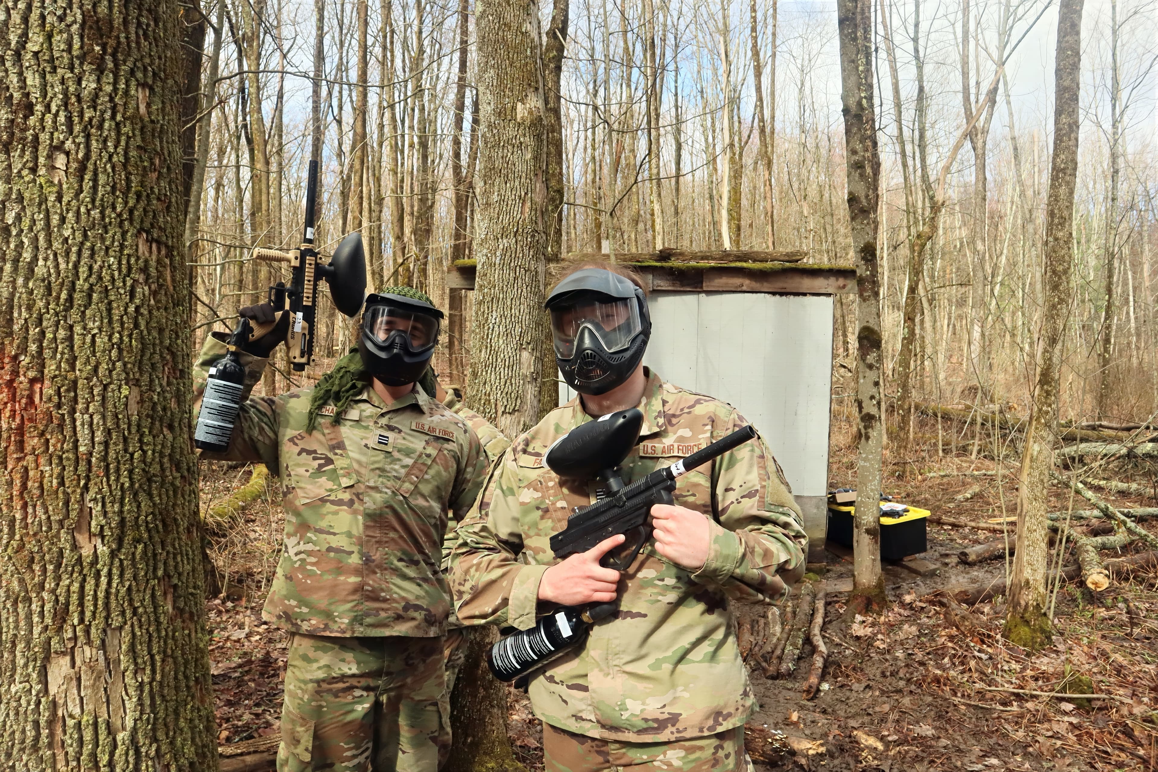 Two cadets with a training marker and a camera pause in the treeline.