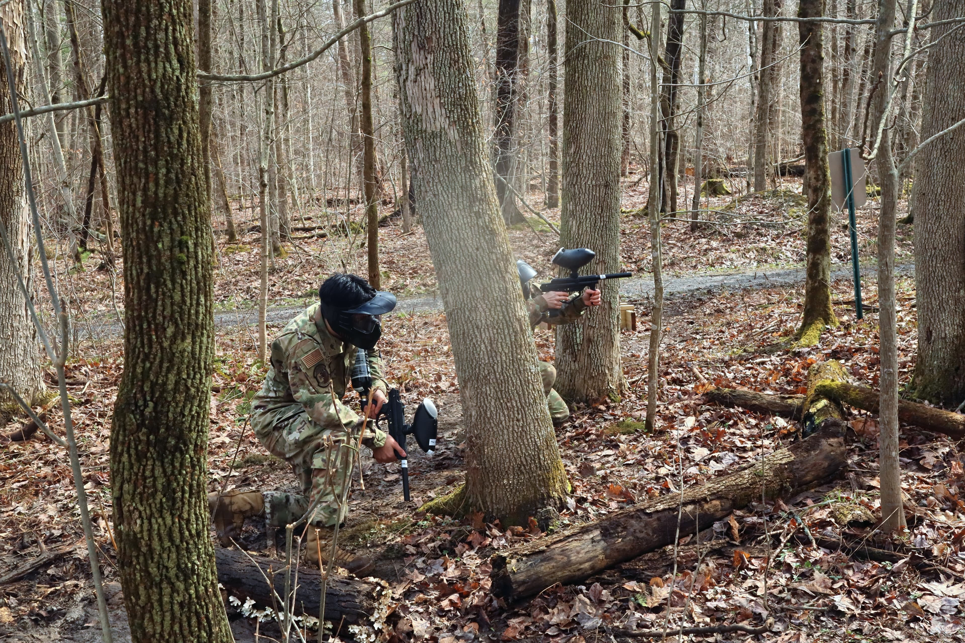 A cadet trains a paintball marker from behind a tree while a teammate observes.