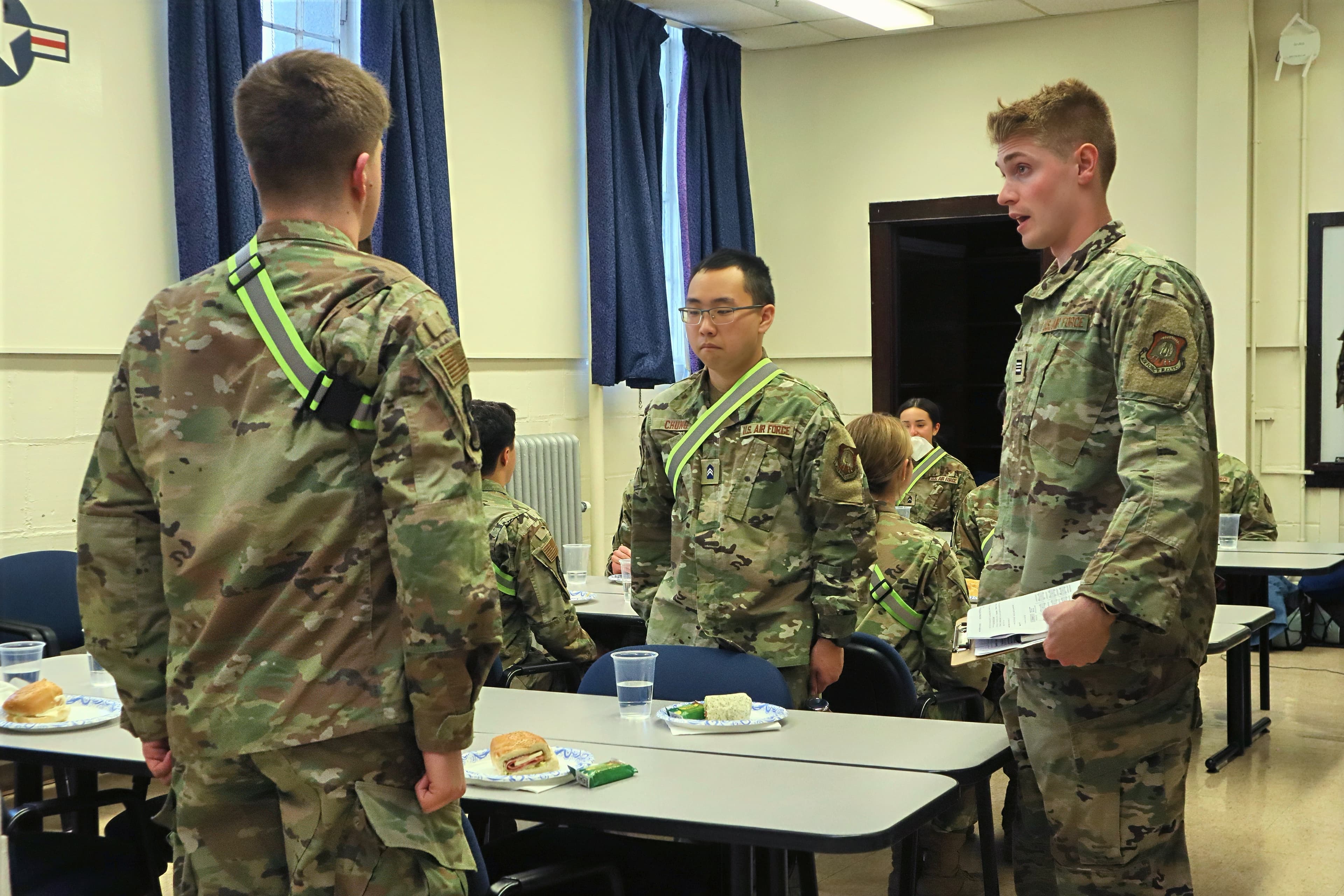 A cadet stands to speak during an in-exercise evaluation while teammates eat lunch.