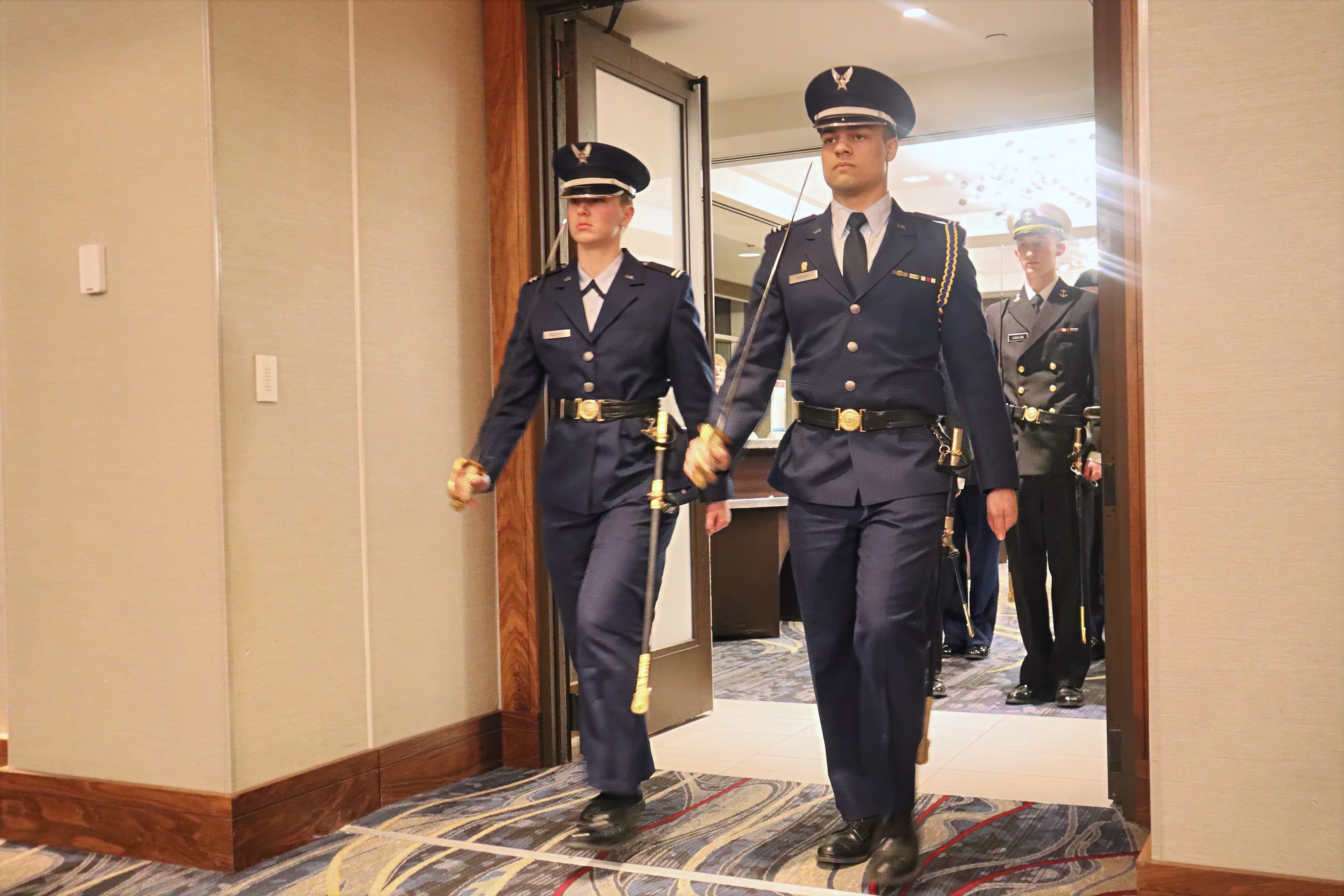 Two color guard members in service dress carry sabers through a doorway at a formal event.