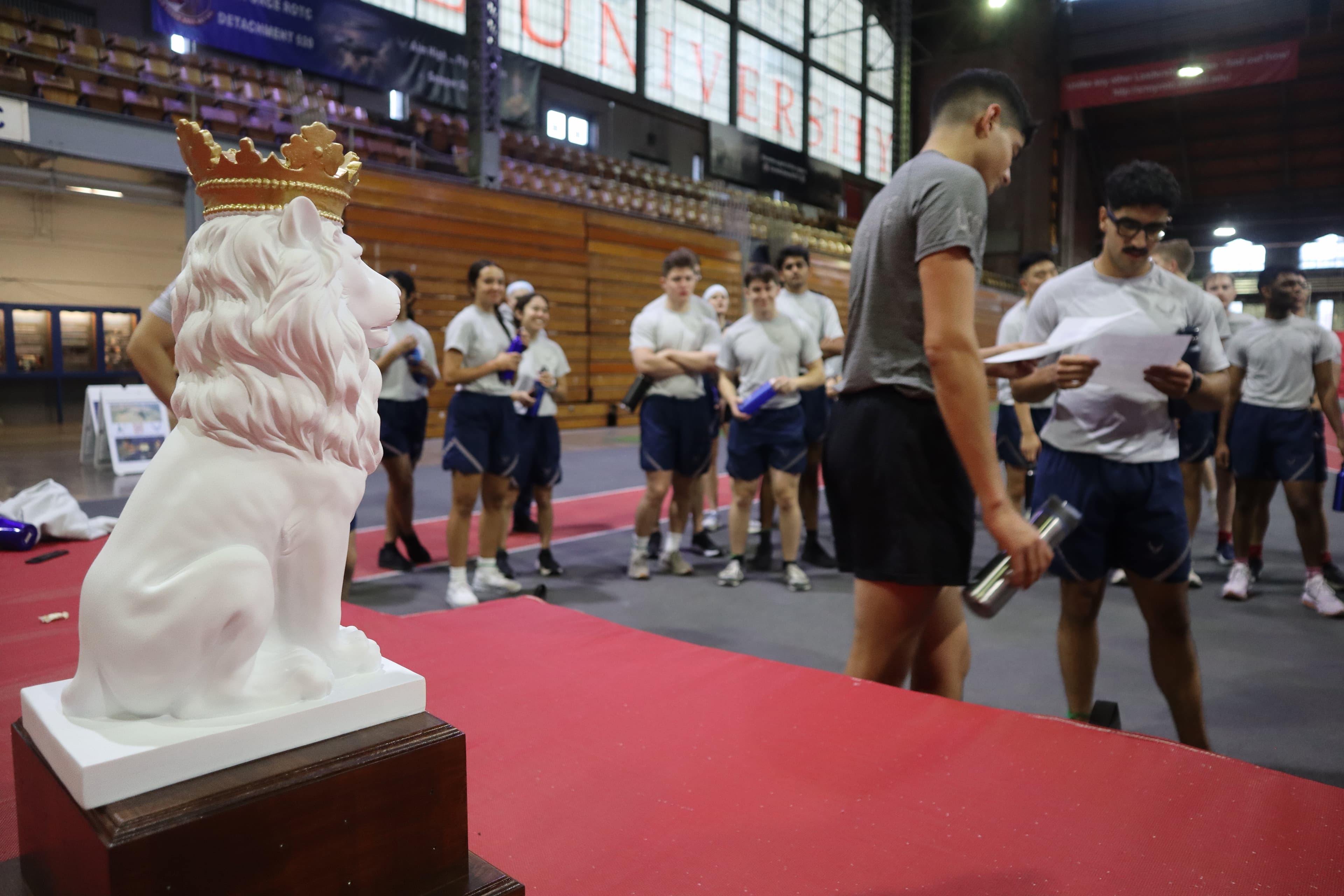 A crowned lion trophy displayed on a red platform in Barton Hall as cadets in physical training gear walk past.
