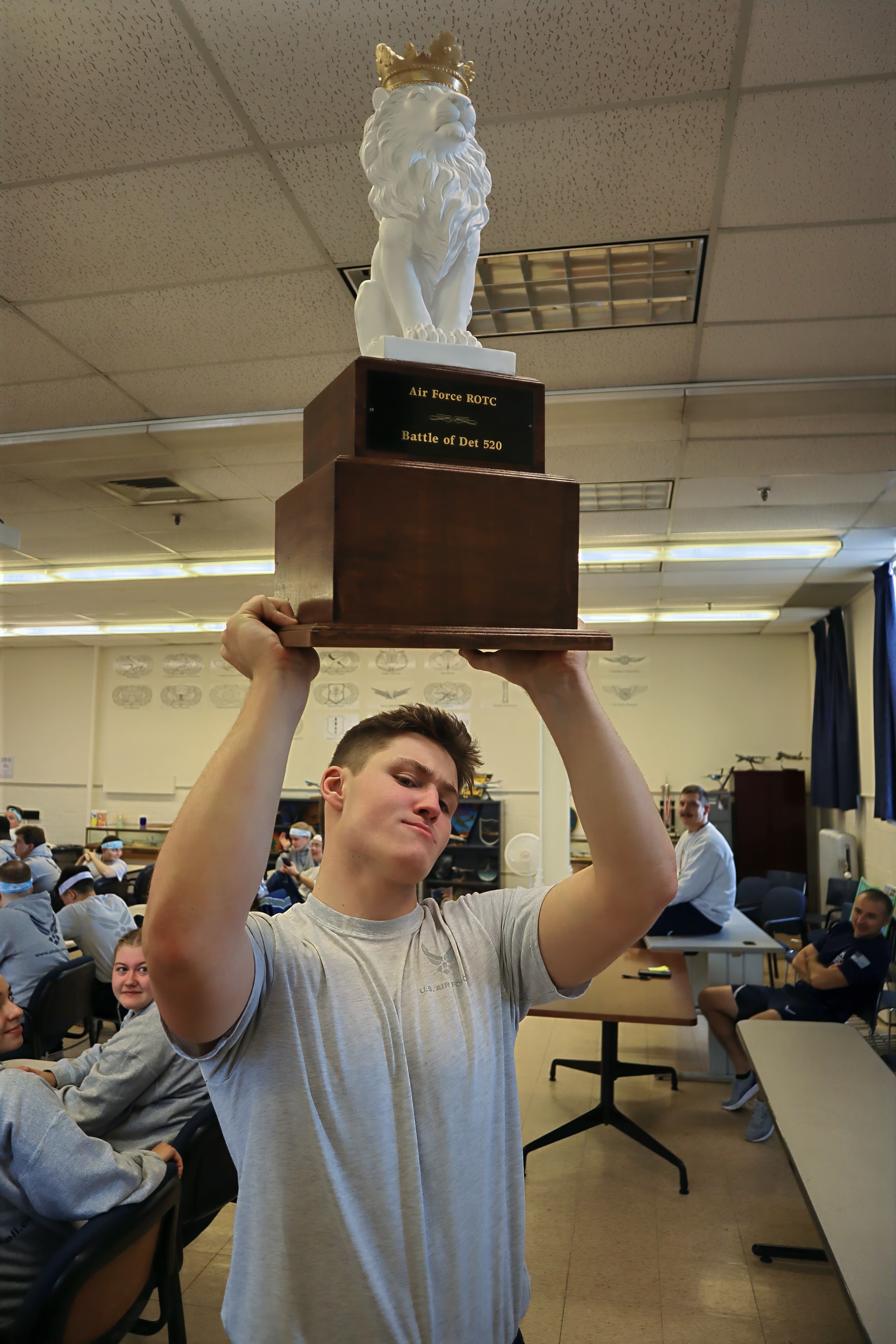 A cadet hoists the lion trophy overhead in a classroom while teammates look on.