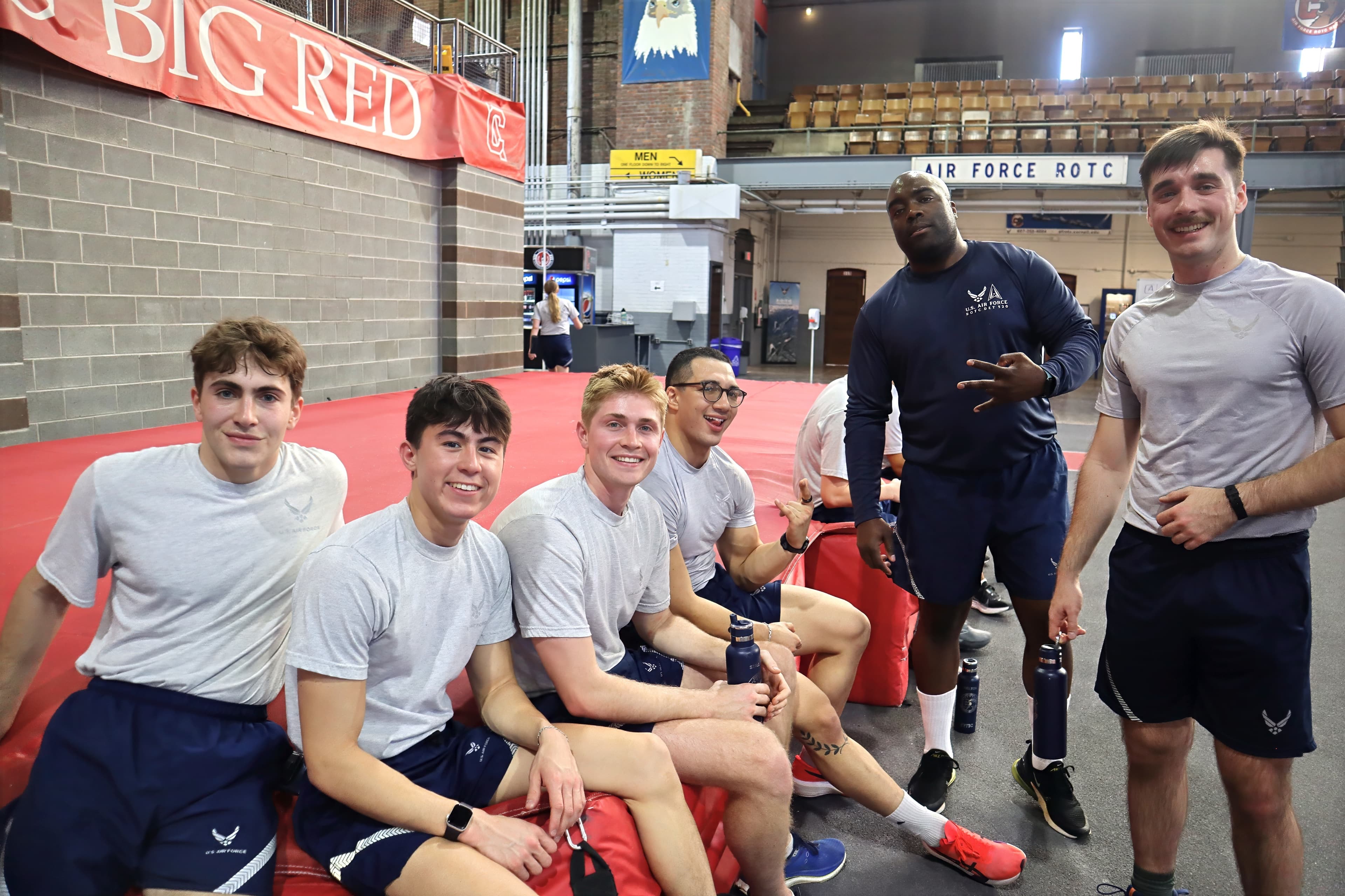 Cadets and cadre in physical training gear rest on red mats under the Air Force ROTC sign after a workout.