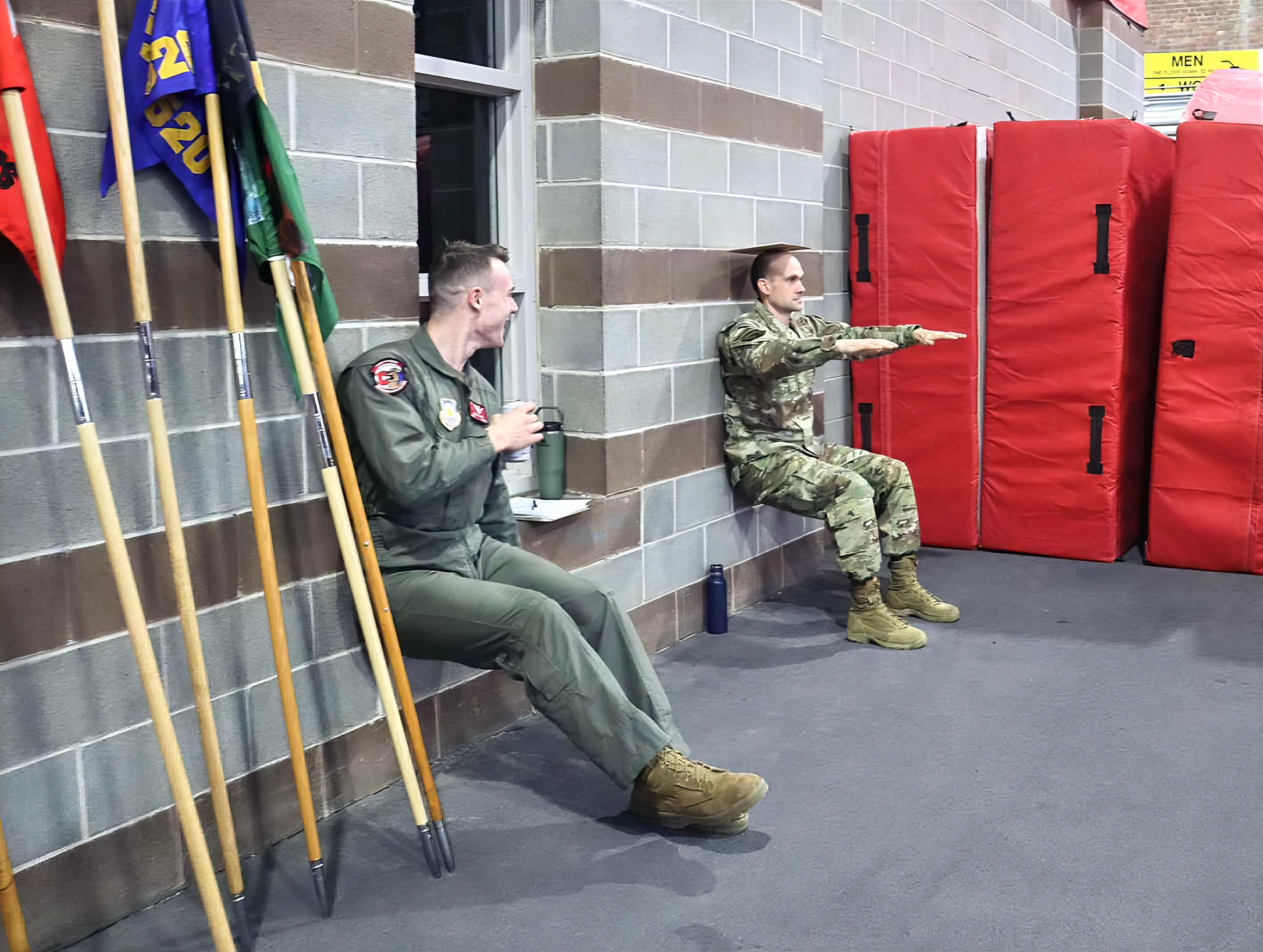 Two flight-suited members hold a wall-sit against the Barton Hall wall, one holding a mug.