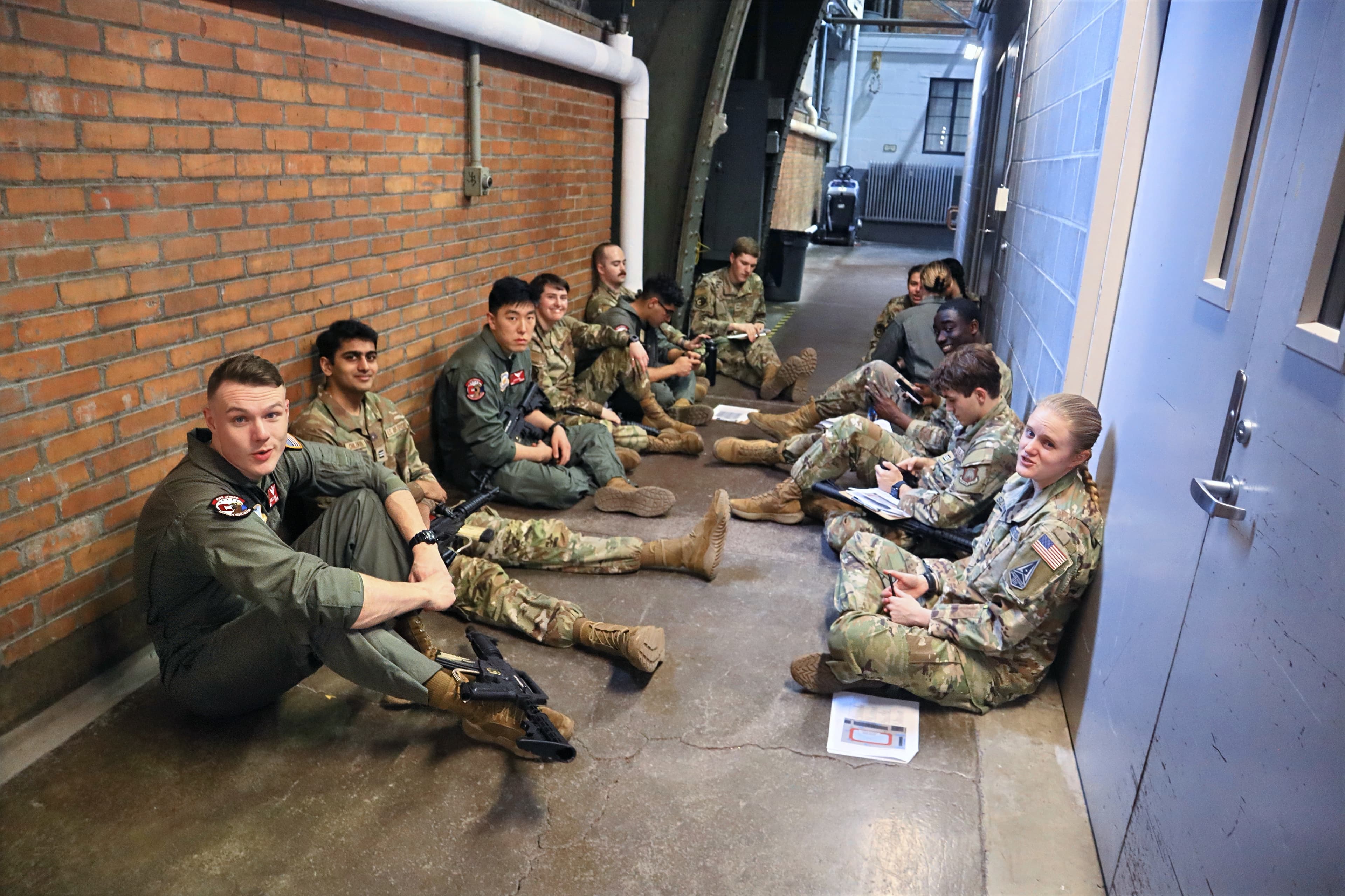 Cadets and a flight-suited member sit in a line against a brick wall pretending to hide.