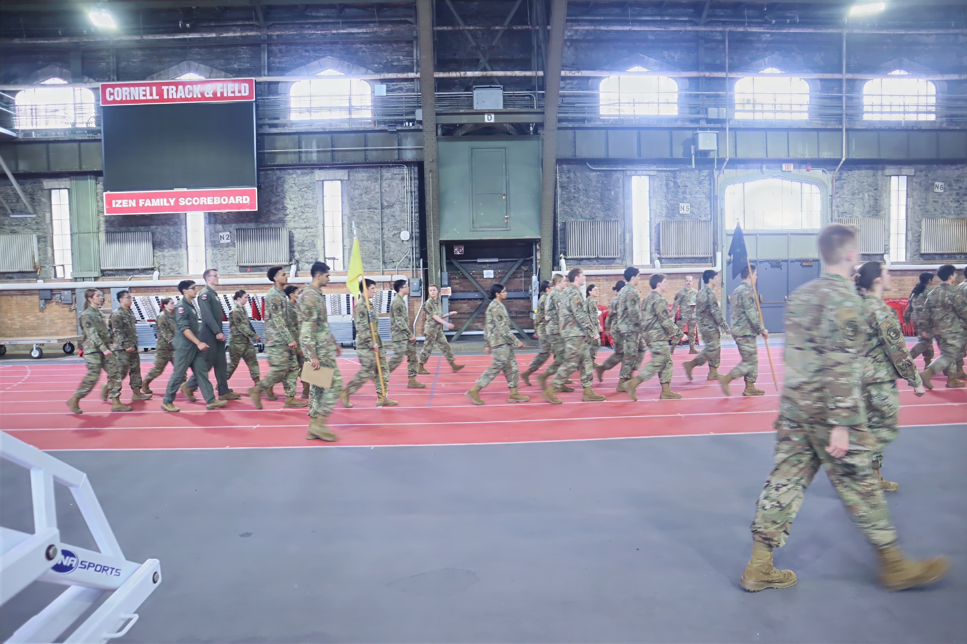 A squadron marches along the track carrying guidons past the scoreboard.