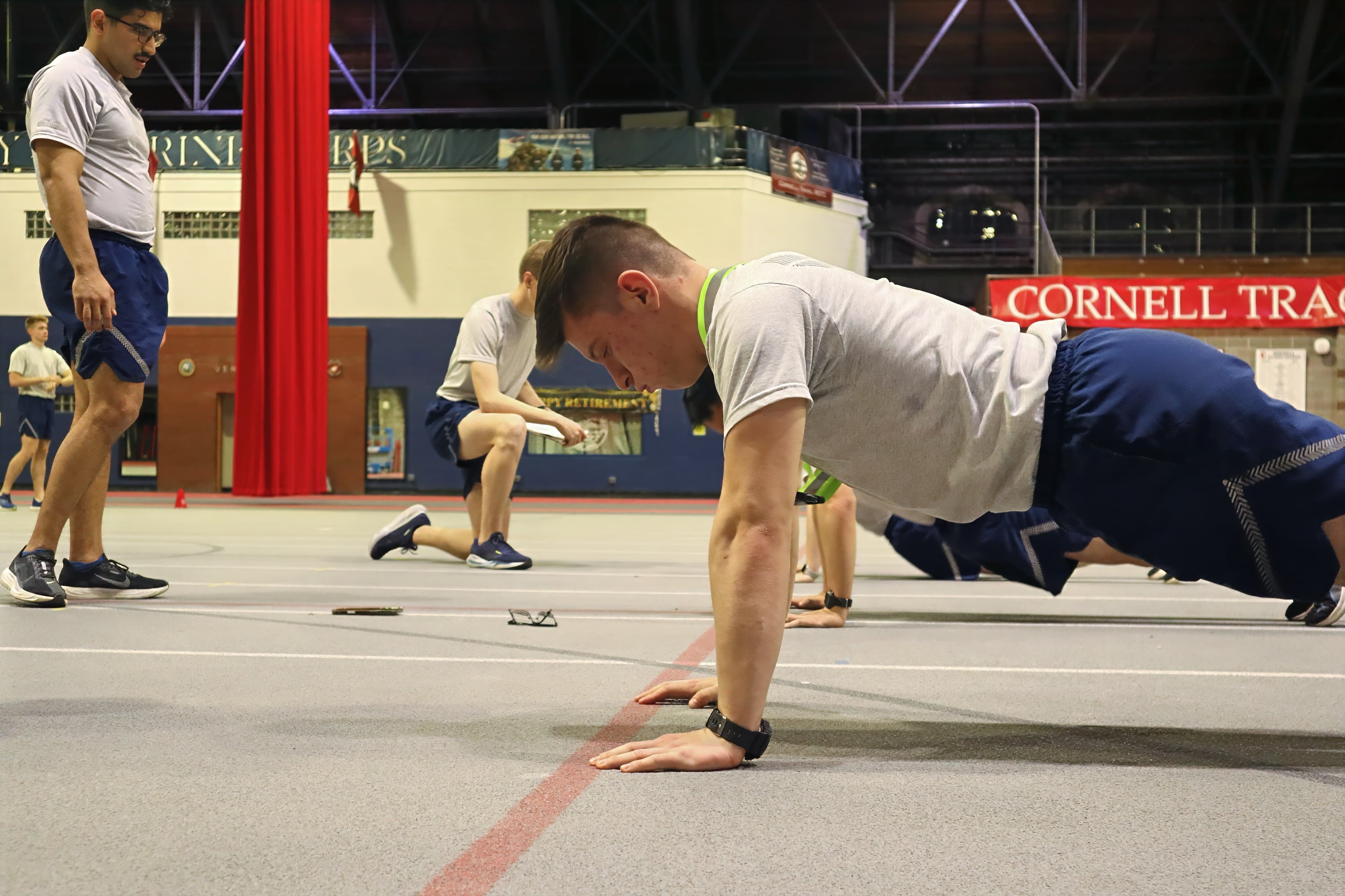 A smiling cadet does pushups on the track beside a red Cornell sign.