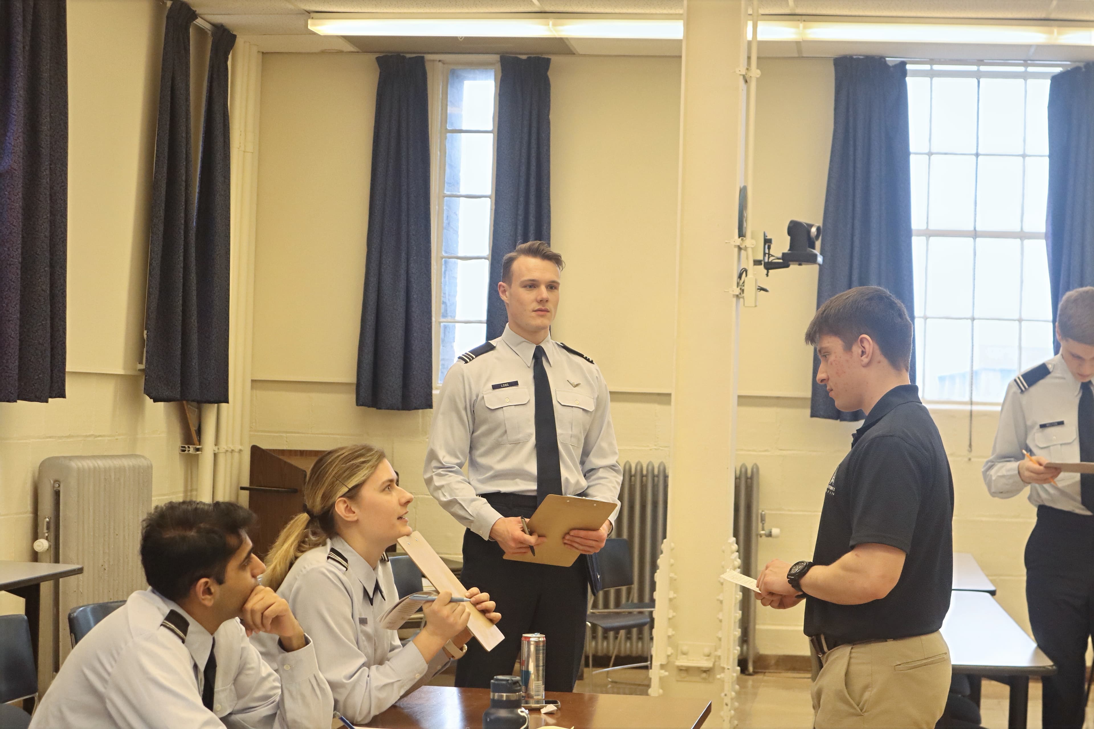 A cadet in civilian clothes reports to two cadets in service dress seated at an evaluation table.