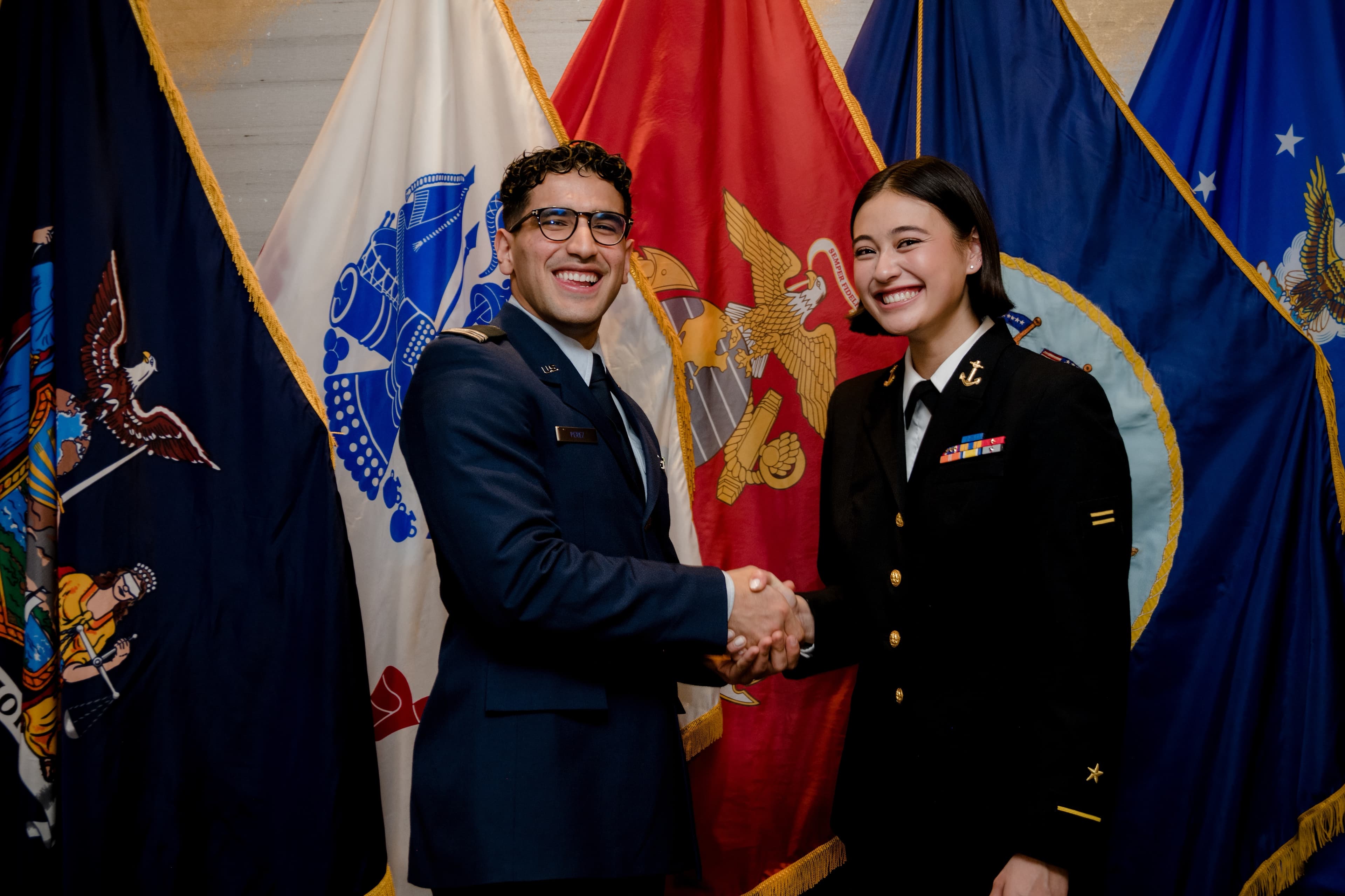 An Air Force cadet and a Navy midshipman shake hands in front of the service flags.