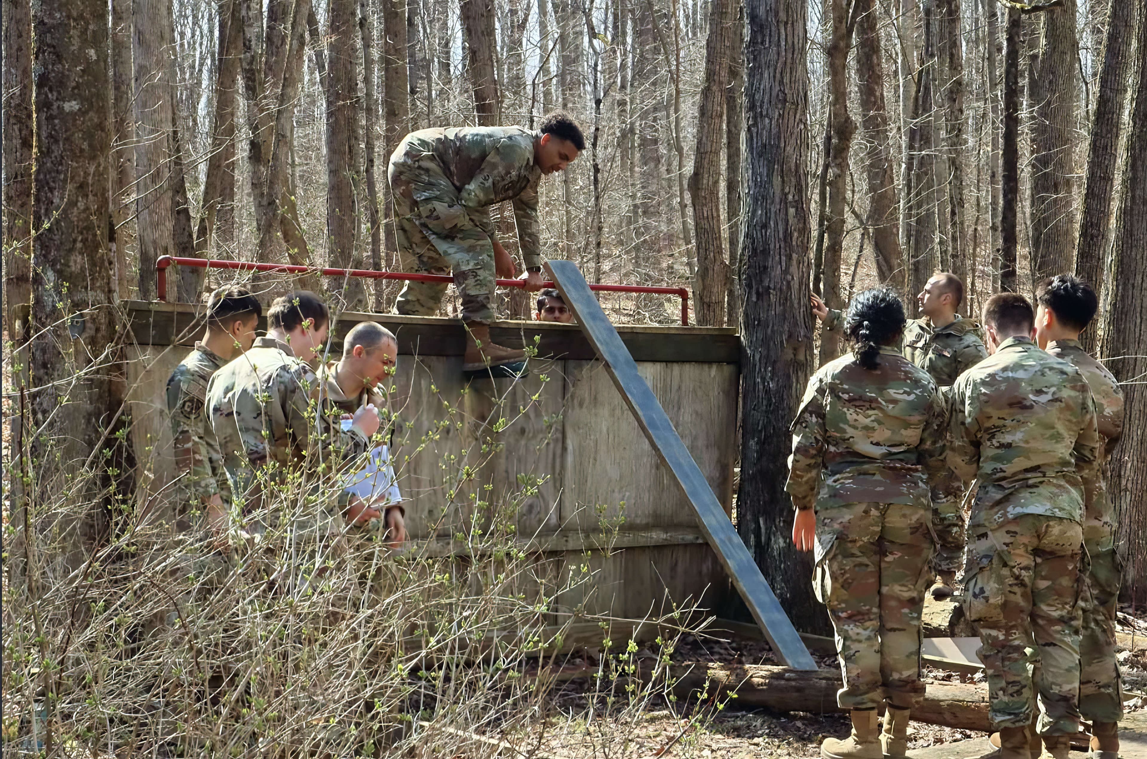 Cadets working together to move a plank across a wooden wall obstacle on the leadership reaction course.