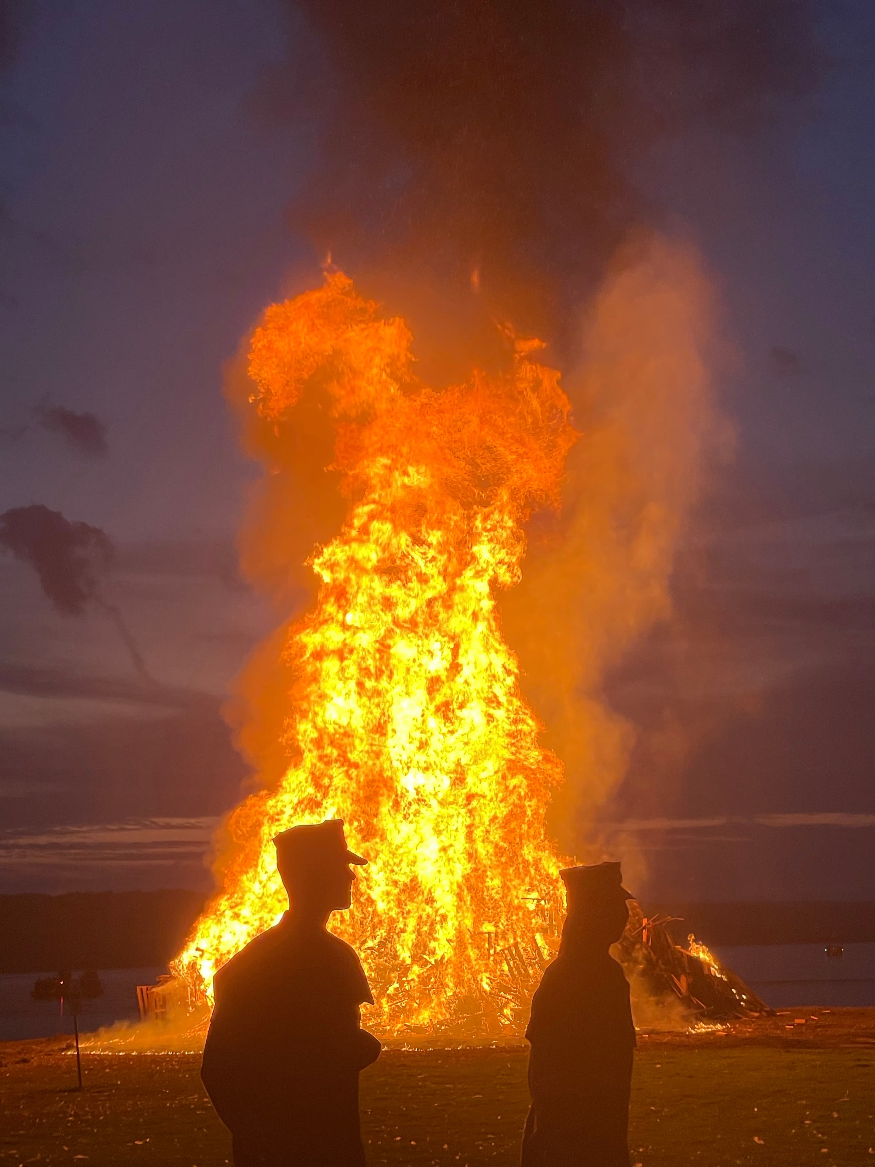 A tall bonfire burns at dusk while two cadets stand silhouetted against the flames.